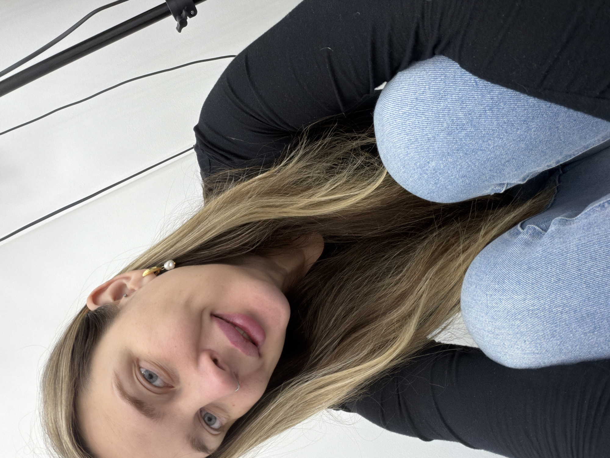 A woman is posing for a selfie. She is wearing a black long-sleeved shirt and light blue jeans. She is blonde with blue eyes. The background is a white wall with a black boom stand in the upper left corner. There is no indication of Moscow, Russia.
