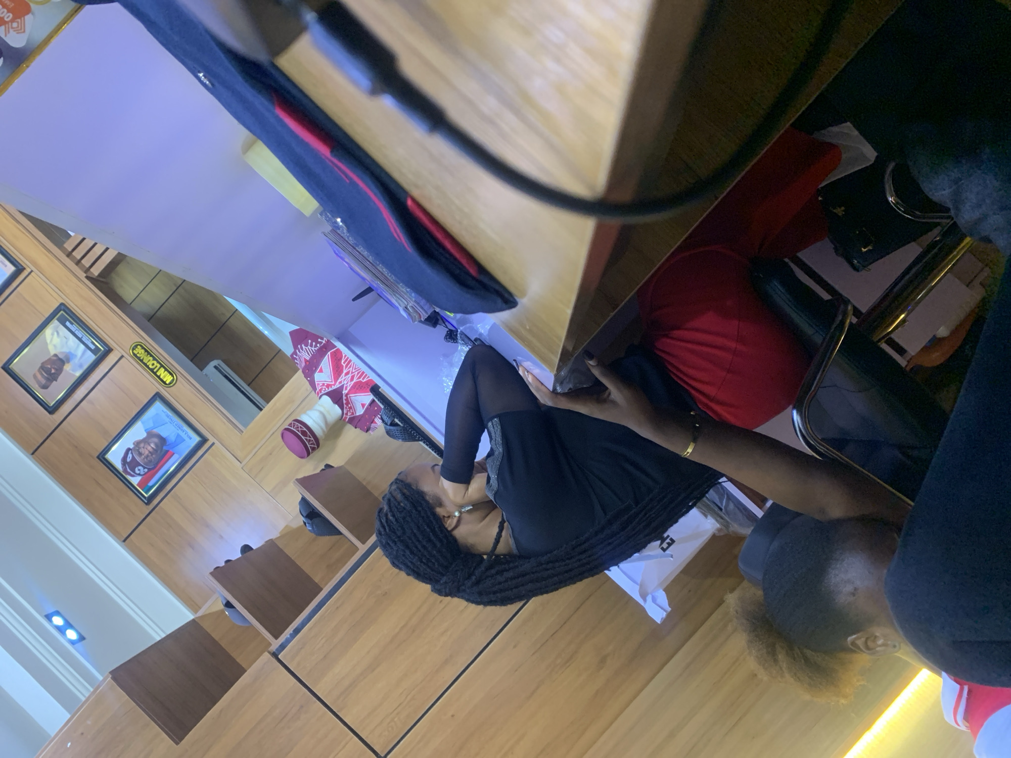 The image depicts an indoor scene, likely a commercial establishment, with wooden paneling on the walls and shelves. In the foreground, a light-colored wooden counter or table is visible, with a black cable extending across it. On the counter, there are stacks of papers and a black and red fabric item.
In the mid-ground, two individuals are present. One person, with dark braided hair, is seated and appears to be resting their head on their hand, with their eyes closed. They are wearing a black top with mesh sleeves and red pants. A gold bracelet is visible on their wrist. To their left, another person is partially visible, with their head down, wearing dark clothing.
In the background, shelves are mounted on the wooden walls. On one shelf, there is a white bag. Another shelf displays framed pictures of men in traditional attire. A yellow sign with the text "MINI LOUNGE" is also visible. The overall lighting suggests an indoor setting, possibly with artificial lights. The scene does not contain any specific elements that would allow for identification of its precise location in Gura Top, Nigeria.