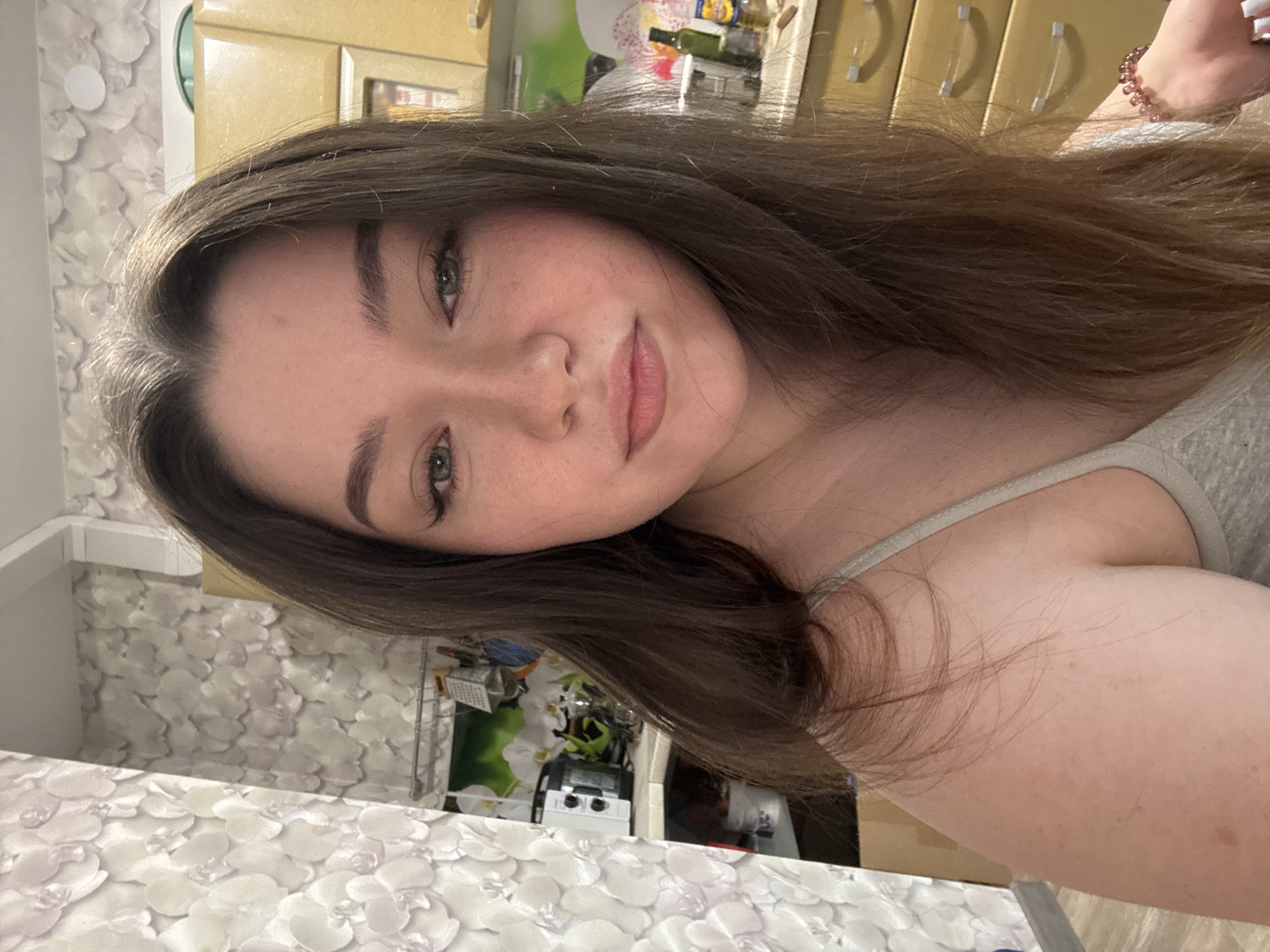 A close-up, head-and-shoulders portrait of a young woman is presented. She has long, dark brown hair, fair skin with freckles, and green eyes. She is wearing a light-colored spaghetti strap top. Her makeup includes eyeliner and mascara. The background appears to be a kitchen area, with yellow cabinets, a backsplash with a floral pattern, and some kitchen appliances visible. A bracelet made of pink beads is visible on her right wrist. The overall lighting suggests an indoor setting, possibly during the daytime. The image quality is clear and sharp.