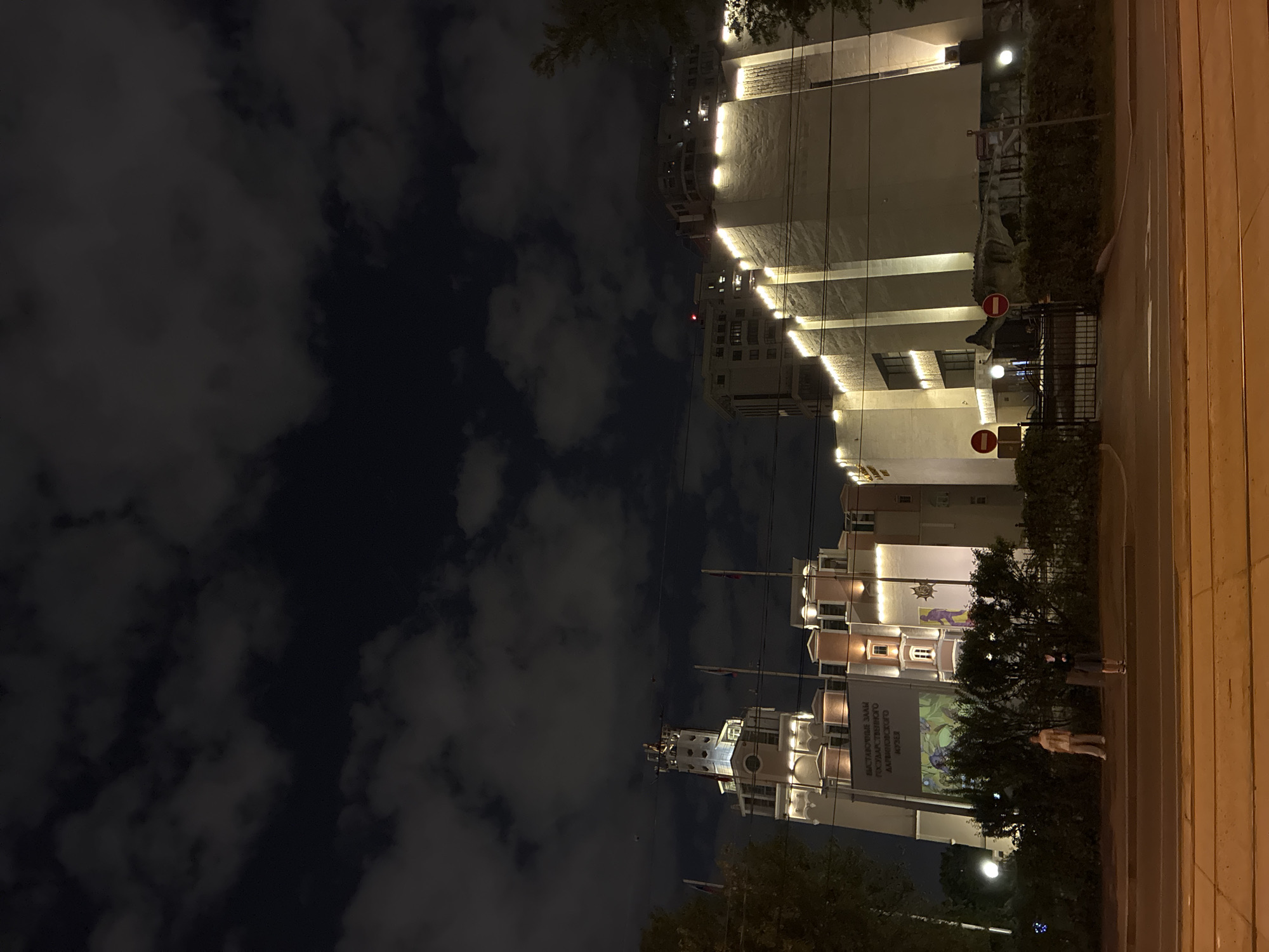 The image shows a night view of a street in Moscow, Russia. In the foreground, there's a sidewalk with a low wall running alongside it. Several multi-story buildings dominate the view, illuminated with white lights. The architectural style is modern with prominent horizontal lines. One of the buildings displays what appears to be a large mural or a sign. People are visible on the sidewalk near the mural. In the background, a cloudy night sky can be seen. There are poles or wires running across the scene, likely for power or communication. Street signs are visible, including "no entry" signs.