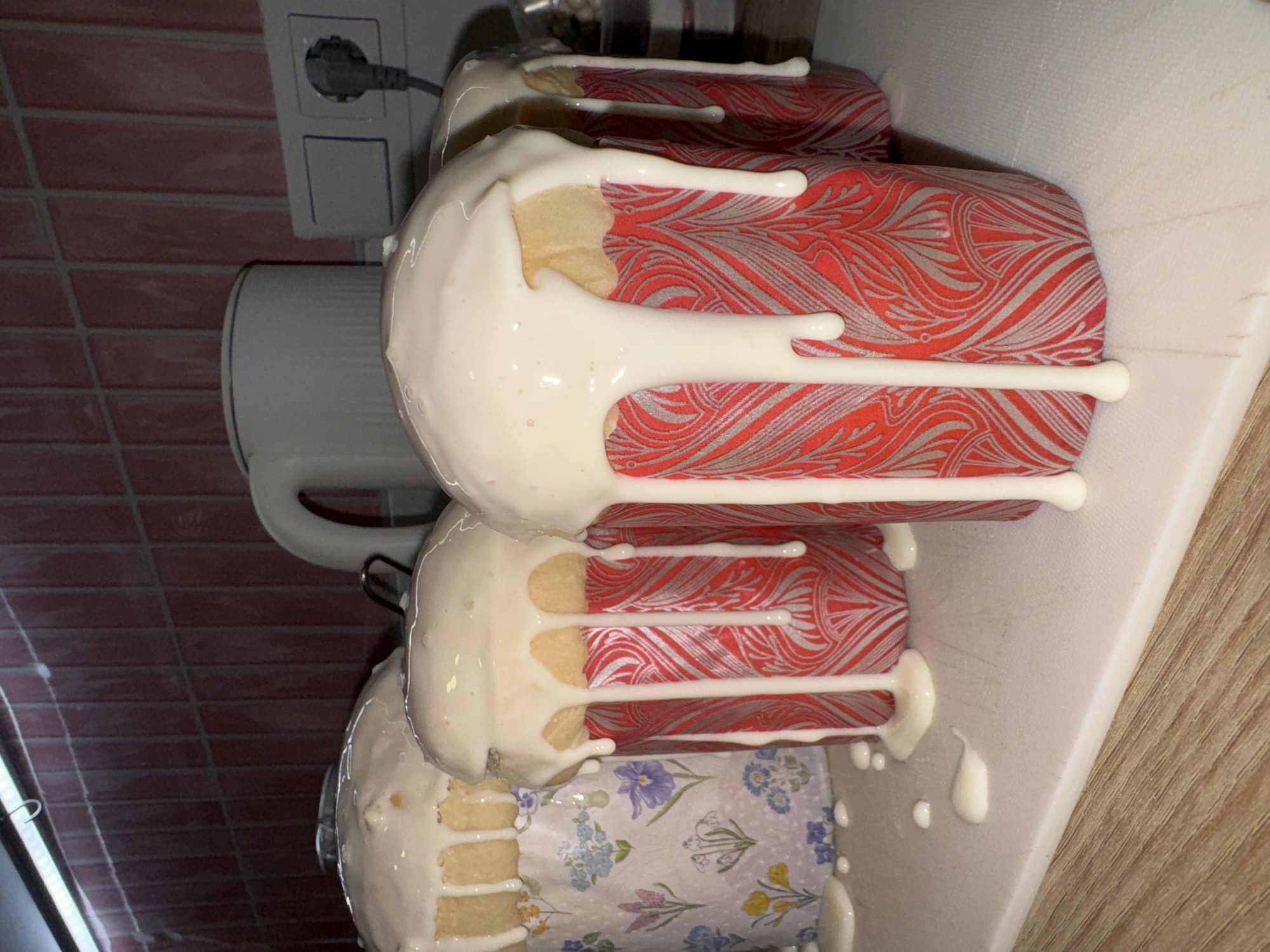 The image shows four decorated Easter cakes (Kulich), glazed with white icing. They are stacked together on a white surface, likely a countertop or tray. The wrappers around the cakes have different designs. One has a red and gray pattern; another has a floral pattern with blue and yellow flowers. A white electric kettle is visible in the background, along with a power outlet on a wall with reddish tiles. The location cannot be determined based on the image alone, and there's no explicit indication of Krasnoyarsk, Russia. The primary focus is on the cakes themselves, their icing, and decorative wrappers. The scene suggests a kitchen setting where the cakes were likely prepared.