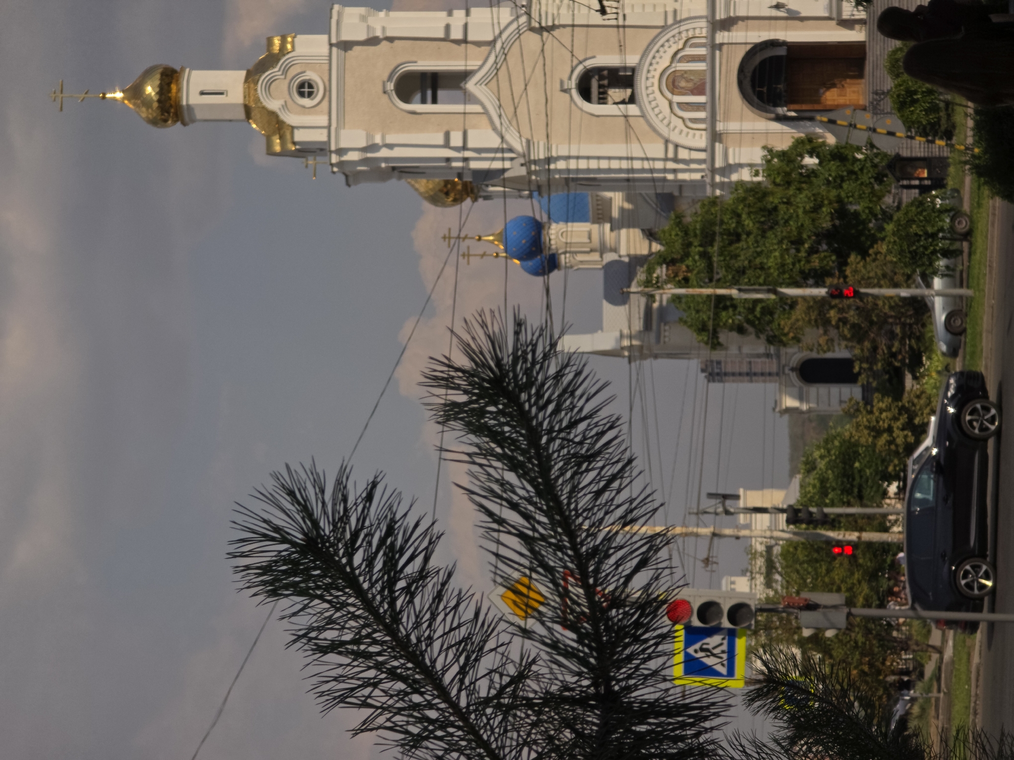 In Rostov-on-Don, Russia, a black SUV is parked on a street with a pedestrian crossing signal displaying a red illuminated man. The traffic light is also red. In the background, a church with multiple domes, some gold and some blue, stands prominently against a cloudy sky. The church has a grand entrance with stairs leading up to it. Green trees are scattered throughout the scene, and power lines crisscross the sky. A branch of a coniferous tree is in the foreground, obscuring part of the view.