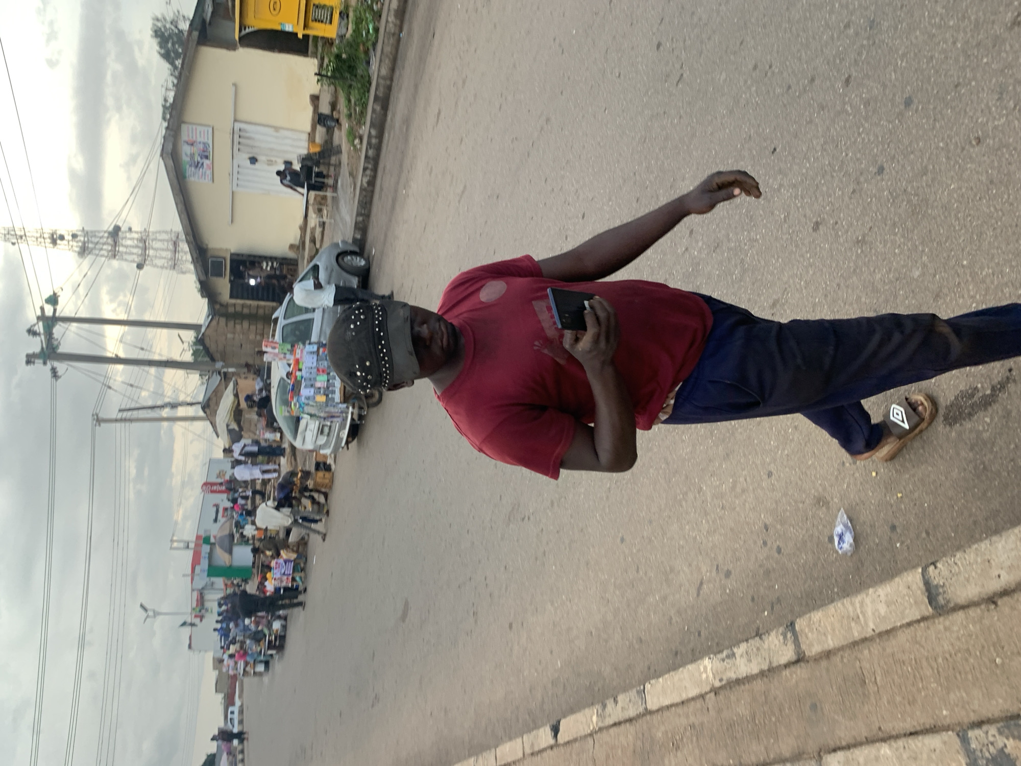 The media file shows a man wearing a red t-shirt and dark pants, holding a smartphone in his right hand. He is standing on a paved street with a sidewalk. In the background, there are buildings, stalls set up along the street, a telecommunications tower, and a yellow vehicle. The scene appears to be a market or a busy street in a town or city. Several other people are visible in the background, some attending to stalls and others walking. There are various goods displayed on the stalls, including what looks like electronic items or accessories. A small, white, crumpled piece of plastic is on the ground near the man's feet. The sky is overcast, suggesting a cloudy day. The lighting indicates it is likely daytime. The overall impression is of a lively, commercial area. The image is oriented vertically.