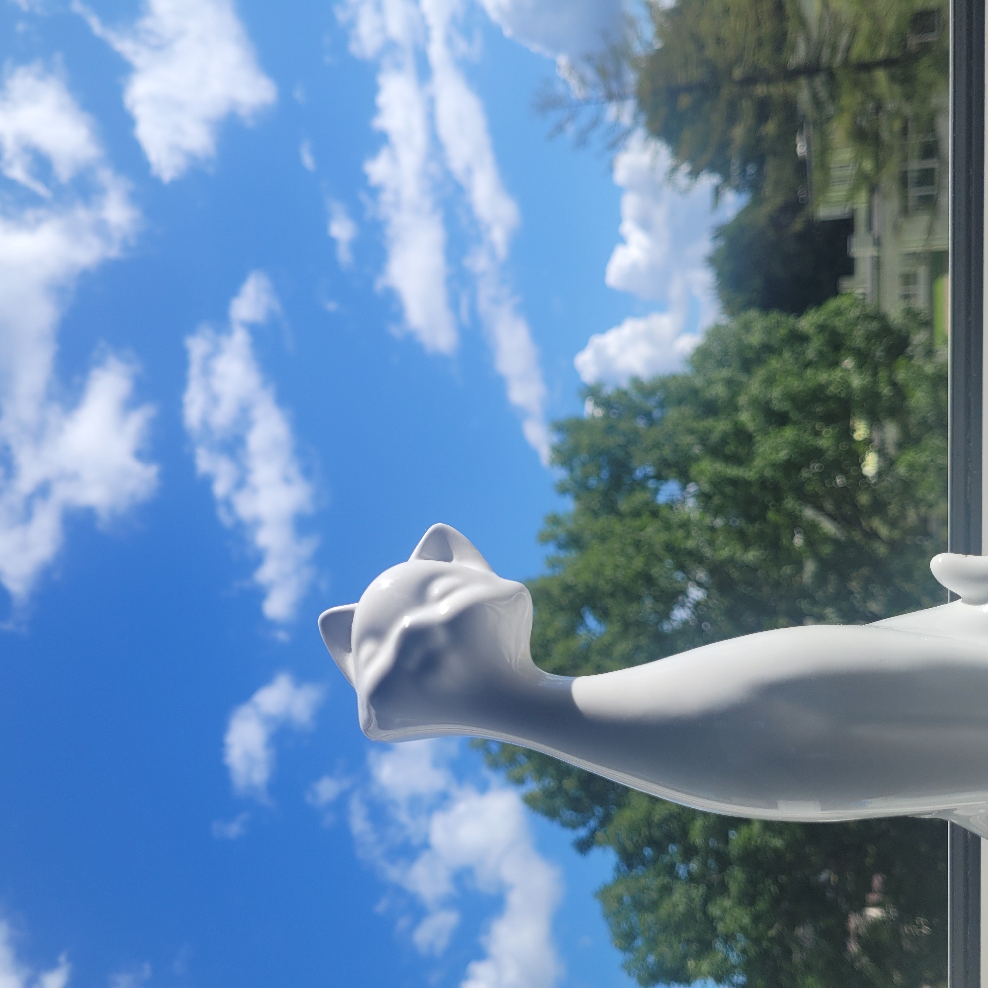 A white ceramic cat figurine is positioned in the foreground, facing left and looking upwards towards a bright blue sky with scattered white clouds. The figurine's head and neck are clearly visible, with its ears pointed and its face subtly sculpted. Behind the figurine, a row of lush green trees forms a dense backdrop. In the distance, beyond the trees, part of a white building with windows is visible. The scene appears to be outdoors, possibly on a windowsill or balcony, with the horizon line marked by a dark horizontal bar in the lower part of the image. The overall impression is one of a serene, sunny day. The specific location in Jūrmala, Latvia cannot be determined from the image alone.