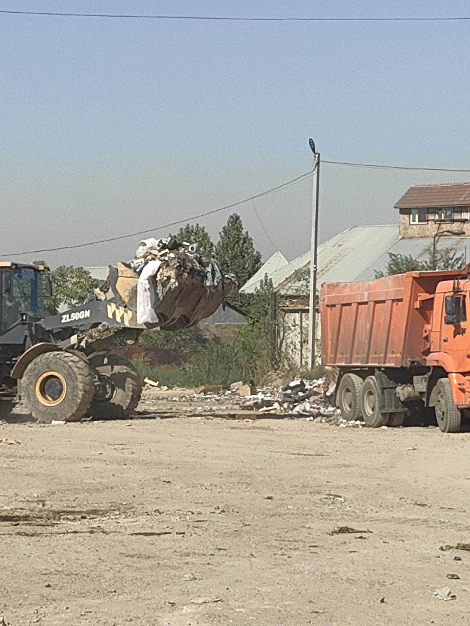 An orange dump truck is parked next to a yellow front-end loader, which is in the process of dumping a large load of garbage into the truck. The scene is outdoors, on a dirt lot. In the background, there are several buildings and trees. The sky is clear and blue. The dump truck has six wheels and appears to be fully loaded. The front-end loader has a large bucket filled with various types of trash, including plastic bags, paper, and other debris. The area around the truck and loader is littered with more garbage. The location is identified as Shymkent, Kazakhstan.