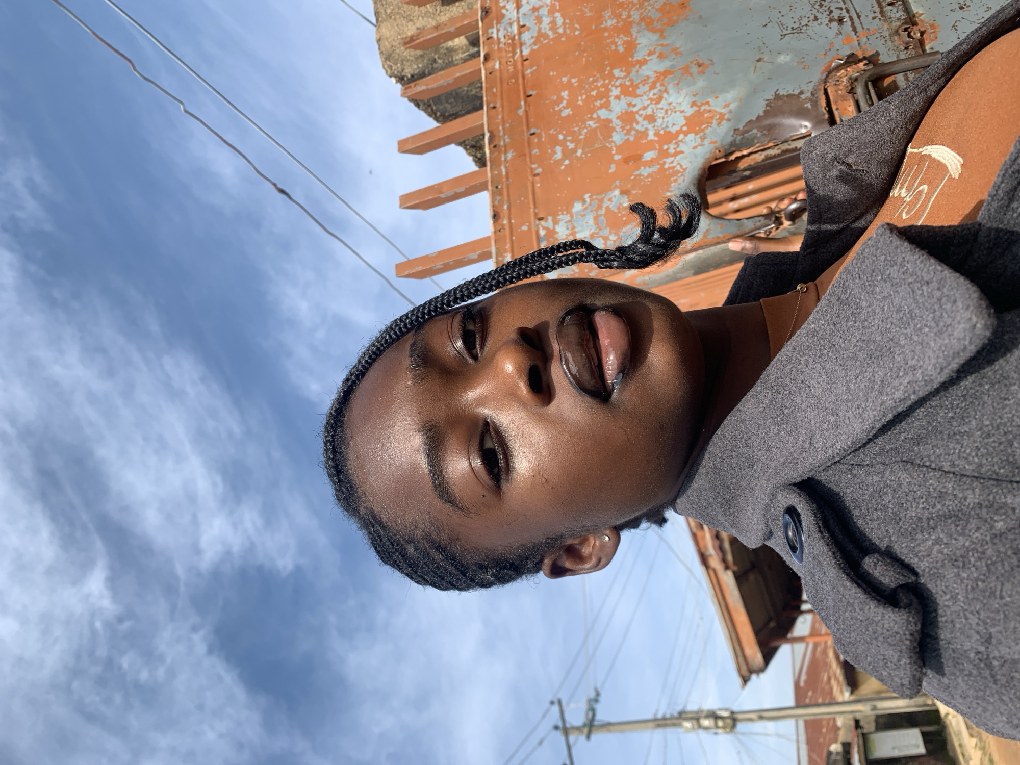 A young Black woman with braided hair and dark skin is pictured from the chest up, outdoors. She wears a grey coat over an orange top, and a thin gold necklace. Her left ear is visible with a small stud earring. She is looking at the camera, with her lips slightly parted and a glossy sheen.
Behind her, the background features a bright blue sky with wispy white clouds. To her right, a large, rusty orange industrial object with vertical slatted elements is visible. The scene is framed by utility poles and wires on the left, and appears to be a street or industrial area. This setting is consistent with the urban environment of Rafin Par, Nigeria, an area known for its bustling street life and industrial presence.