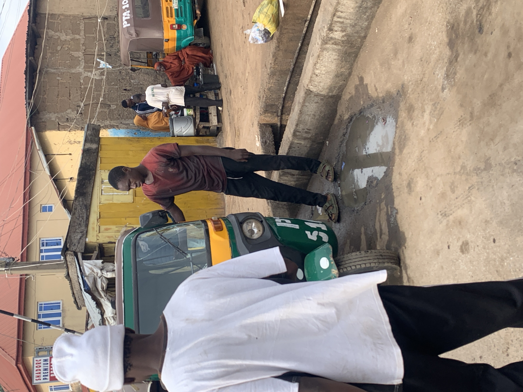 The image captures a street scene in Gura Top, Nigeria. In the foreground, a person wearing a white beanie and a white t-shirt faces away from the camera, partially obscuring a green and yellow auto-rickshaw. To the right of the auto-rickshaw, a young man in a maroon t-shirt and black pants stands with his hands on his hips. Behind him, several other individuals are visible: a woman in a bright orange headscarf and dress is seated, and another person in a white t-shirt stands near a bucket. Further in the background, a yellow building with blue-trimmed windows is visible, along with a brown and green auto-rickshaw with "PTA-105" on its side. The ground is unpaved and appears to be dusty, with a puddle of water near the curb. The overall impression is that of a typical street corner in a bustling Nigerian city.