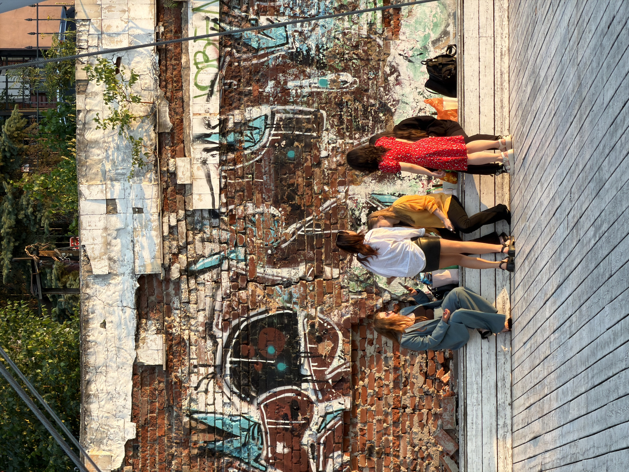 The image depicts a group of five young women socializing on a wooden deck in front of a graffiti-covered brick wall, located near the Kreml building in Kazan, Russia. The women are seated on a white wooden bench. The woman on the far left is wearing a teal suit. Next to her, a woman in a white long-sleeved shirt and black skirt is visible. To her right, a woman in a yellow top and black pants is seated. Another woman in a red floral dress stands to the right of the woman in yellow. The fifth woman, on the far right, is partially obscured and appears to be wearing dark clothing. Several plastic cups and bags are placed on the bench. The brick wall behind them is adorned with colorful graffiti, and some of the plaster on top of the wall is chipped. Green foliage is visible above the wall, and a wire cable stretches across the top of the frame. The lighting suggests it is late afternoon.