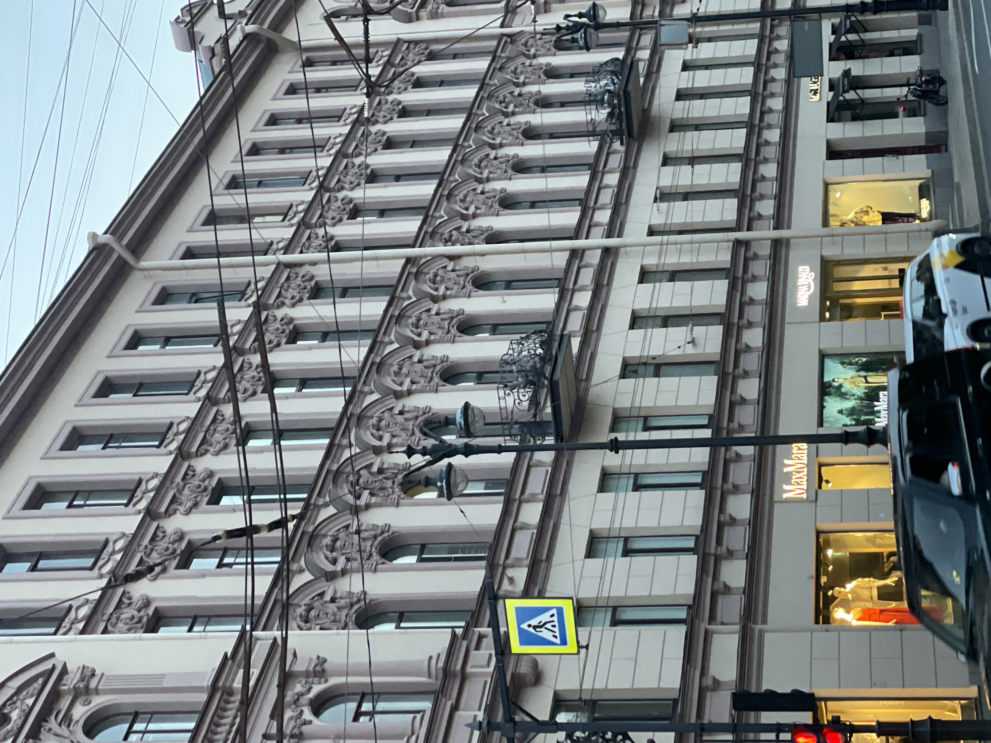 The photograph depicts a multi-story building in Saint Petersburg, Russia. The building has ornate architectural details and evenly spaced windows. In the lower portion of the frame, a "pedestrian crossing" sign in blue and yellow is visible. The words "MaxMara" are seen vertically on the side of the building. Showcases of clothing are seen at the bottom of the frame. A car is seen to the right of the showcases, its reflection is also visible. Electrical wires are visible extending throughout the front of the building. Street lights can be seen on the pole. The sky is overcast.