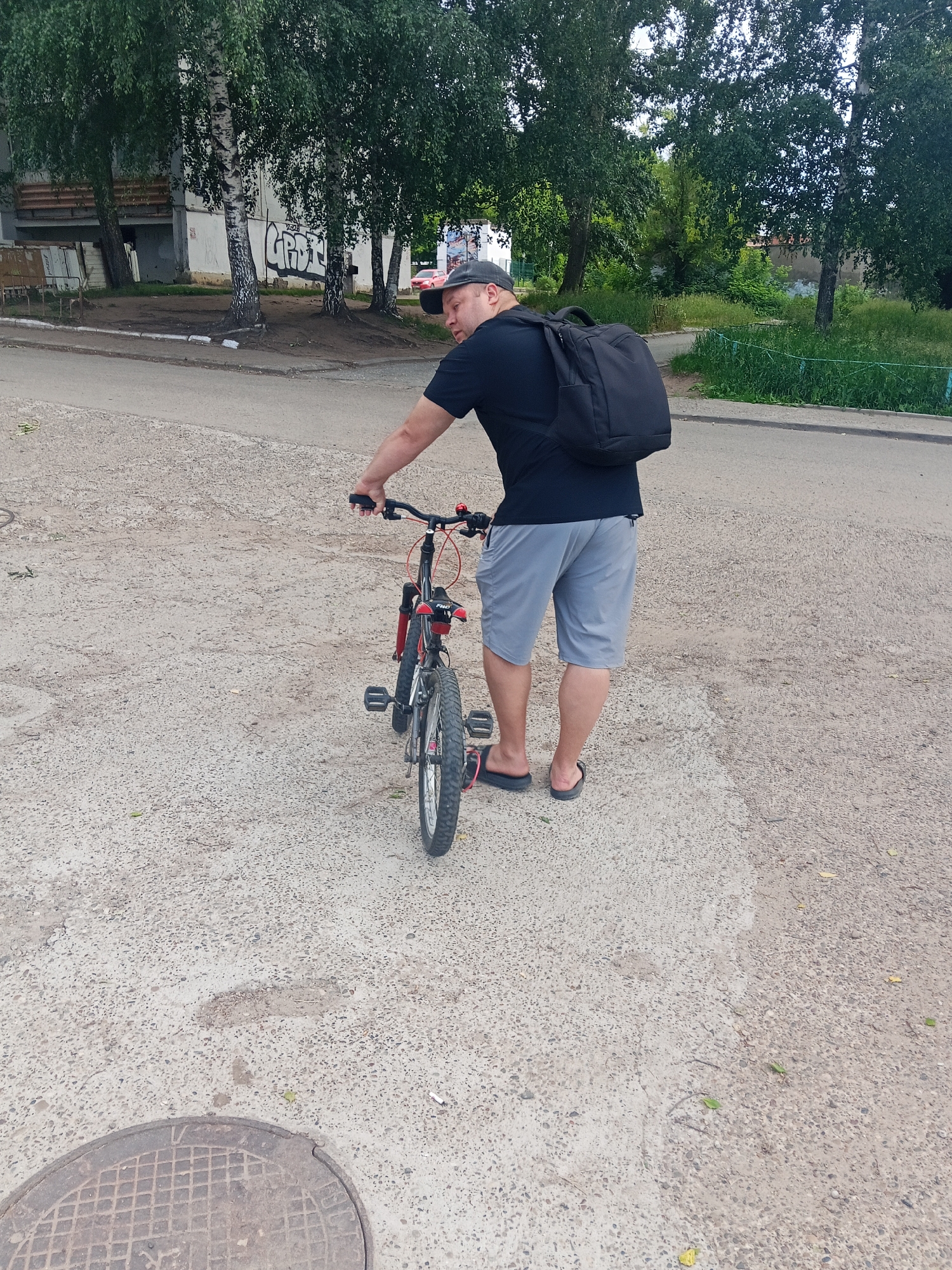 A man wearing a black t-shirt, grey shorts, a dark baseball cap, and black flip-flops stands next to a red and black bicycle on a concrete road in Chaikovsky, Russia. He has a black backpack on. The road is bordered by trees and a building with graffiti. A manhole cover is visible on the left side of the frame. The sky is overcast.
