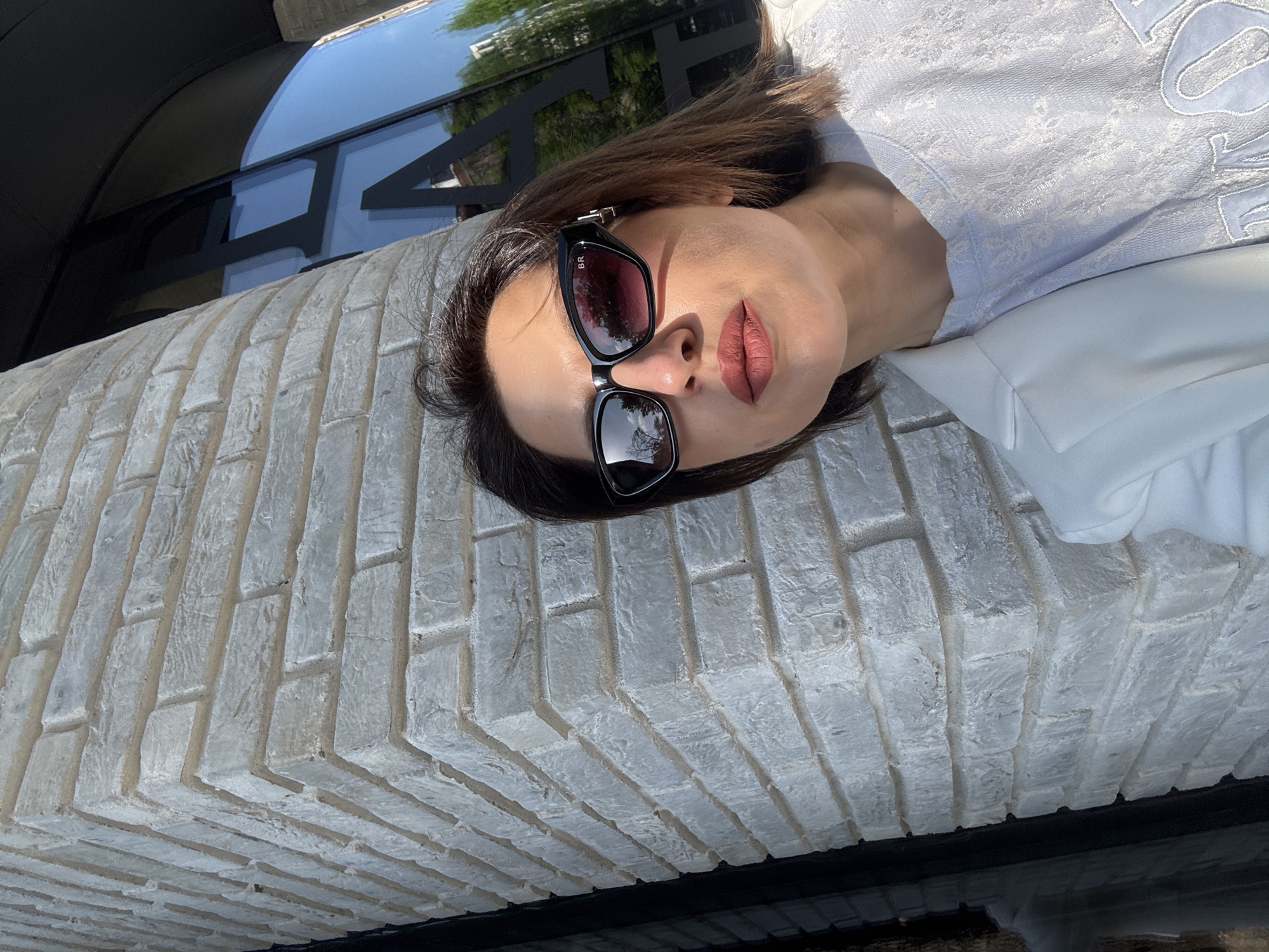 A woman wearing sunglasses is lying on a ledge made of light grey bricks. She is wearing a white blouse with a textured, embroidered design. In the background, there is a reflective window with the name "CHANEL" displayed on it. The location is Sofia, Bulgaria.