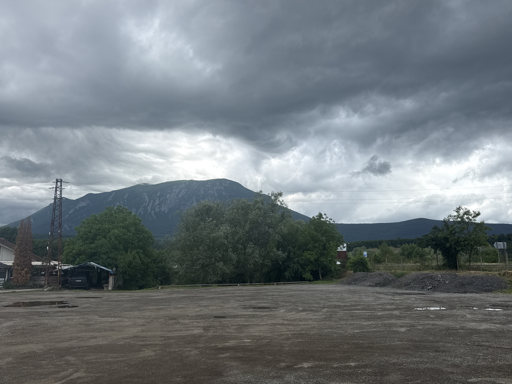 This image presents a wide view of an outdoor scene in Kravoder, Bulgaria, characterized by a dramatic, heavily overcast sky. The foreground features a large, empty, unpaved dirt and gravel lot with scattered puddles, indicating recent rain.
In the mid-ground, a mix of structures and dense vegetation separates the open lot from the mountainous background. On the far left, a section of a building is visible next to a tall, slender evergreen tree. Adjacent to this, a prominent metal lattice transmission tower stands, connected by power lines that stretch horizontally across the scene. To the right of the tower is a makeshift shelter or shed with a light-colored, possibly corrugated, roof, under which a dark-colored car is parked. A thick line of large, green deciduous trees forms a natural barrier, with a simple wooden fence partially visible behind them. Further to the right, two mounds of dark gravel or aggregate material are piled on the edge of the lot.
The background is dominated by an impressive mountain range. A large, steep mountain with a rocky face rises prominently in the left-center, while lower, forested hills extend across the right side of the frame. The sky is filled with voluminous, dark grey clouds, creating a subdued, pre-storm or post-storm atmosphere. The light is diffused, suggesting it is daytime despite the heavy cloud cover. No people are visible, and no specific activity is taking place, giving the scene a quiet and somewhat desolate impression. No legible text is discernible in the image.