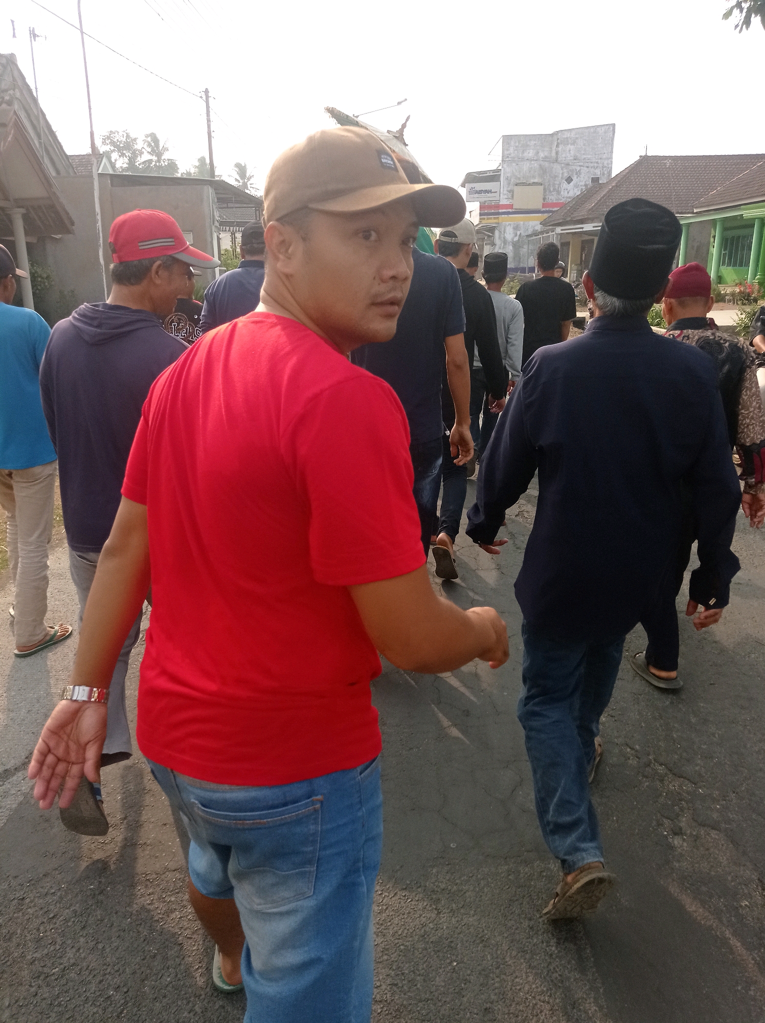 The image depicts a group of people walking on a paved road in what appears to be a residential area. In the foreground, a man in a red t-shirt and blue denim shorts is looking over his left shoulder. He is wearing a brown baseball cap and a wristwatch on his left wrist. Behind him, several other individuals are walking away from the camera, also on the road. They are dressed in various casual and traditional clothing, including t-shirts, hoodies, and some individuals are wearing caps or head coverings. The environment suggests a sunny day with buildings and trees visible in the background. The road is asphalt with visible cracks. The presence of buildings and a general streetscape indicates an urban or semi-urban setting.