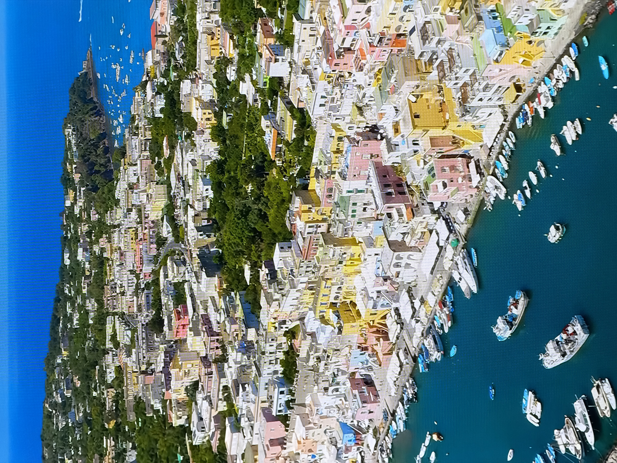The image depicts a coastal town with colorful buildings cascading down a hillside towards a harbor filled with numerous boats. The architecture is dense, with buildings of varying heights and hues, including pink, yellow, white, and blue. Green trees are interspersed among the buildings, and the ocean is a deep blue. The scene appears to be a popular tourist destination, given the presence of many leisure boats and the picturesque setting. This description is unrelated to Chirchik, Uzbekistan.