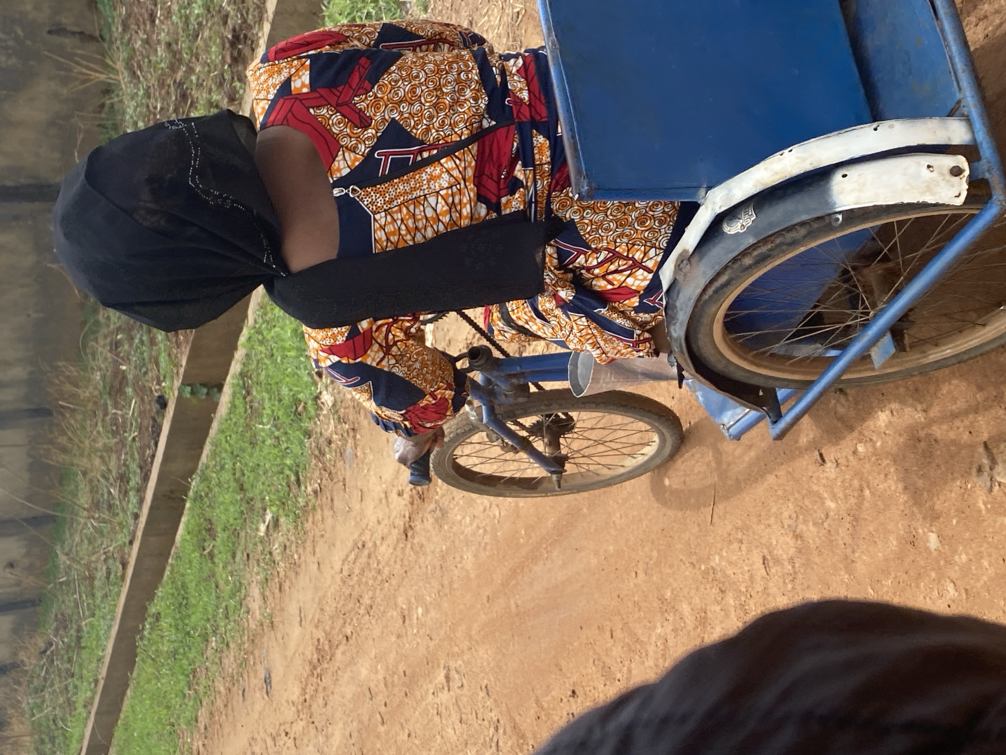 An eye-level, slightly downward-angled shot captures an individual on a reddish-brown dirt path in Maigizo, Nigeria, under natural daylight. The person, viewed from behind, wears a black head covering and a vibrant, patterned dress or top in shades of red, orange, and blue with geometric and swirl designs, cinched at the waist by a black band. They are bent forward, seemingly operating or attending to a blue three-wheeled vehicle, which appears to be a tricycle modified for transport or cargo.
The vehicle features a blue metal frame, two rear wheels with white fenders, and a single front wheel. A blue box-like structure is visible at the rear of the tricycle. The person's hands are near the handlebars or front assembly of the vehicle. To the left of the dirt path, a concrete strip or small ditch separates the path from an area covered in green grass and some sparse vegetation. The overall setting suggests a rural or semi-urban outdoor environment. No discernible text is visible on the clothing or vehicle, and the person's expression is not visible.