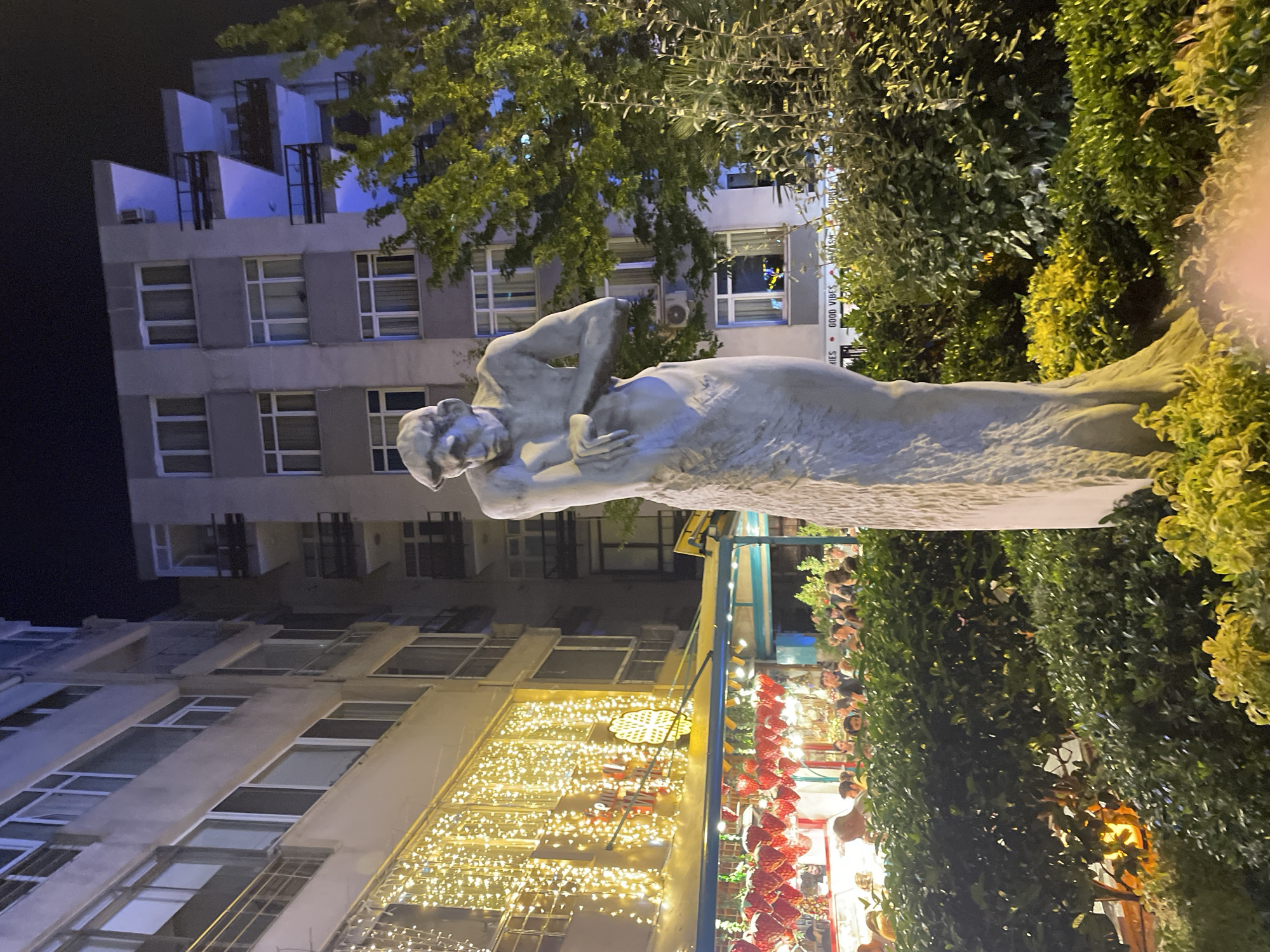 This photograph captures an outdoor scene at night in the city of Kadıköy, Turkey, likely in a commercial or entertainment district. A tall, pale sculpture of a pensive male figure stands prominently in the foreground, its texture rough and weathered. Behind the sculpture, a building with multiple floors and balconies is visible, some illuminated with blue light, suggesting an urban setting with residential or commercial spaces. To the left, a storefront or restaurant is adorned with a cascade of warm, twinkling string lights, creating a festive atmosphere. Beneath this illuminated facade, an awning is decorated with numerous red, heart-shaped objects, possibly ornaments or merchandise. A crowd of people is gathered under the awning and further back, engaged in various activities, though their individual features are indistinct due to the low light and distance. Lush green foliage, including bushes and trees, surrounds the sculpture and the outdoor seating area, indicating landscaping within the urban environment. The overall impression is of a lively evening scene in a city, blending art, commerce, and social gathering.