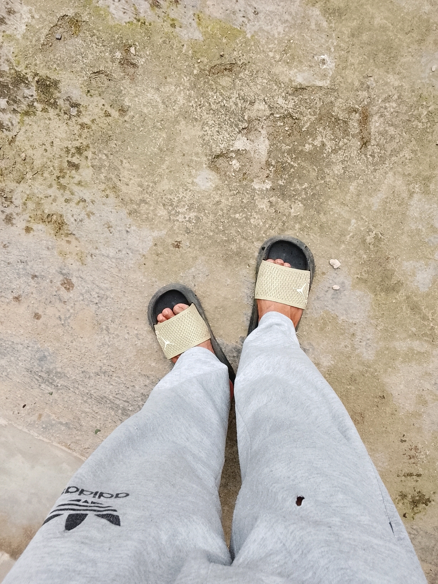 A top-down, medium shot captures a person's legs and feet standing on a textured, concrete surface. The concrete appears weathered and uneven, with patches of discoloration and small stones or debris scattered across it. The person is wearing light gray sweatpants with a black Adidas logo and three stripes visible on the left pant leg. On each foot, the person wears a black slide sandal with a light beige or tan strap, possibly made of a mesh-like material. A small, faded logo, possibly a Jumpman symbol, is visible on the strap of the right sandal. The person's toes are visible through the straps of the sandals. There is a small, dark hole or stain on the right pant leg. The background is predominantly this textured concrete, suggesting an outdoor environment. The scene is devoid of any recognizable landmarks or specific architectural details that would definitively place it in Padang Dalom, Indonesia.
