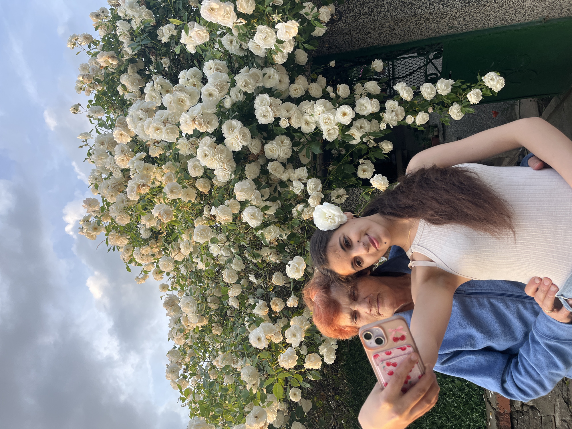 An outdoor selfie captures two women, likely family, against a backdrop of abundant white roses in Lozarevo, Bulgaria. The older woman, with short, reddish-orange hair and wearing a blue zippered jacket, holds a smartphone to take the picture. The phone is an iPhone with dual cameras, encased in a clear cover adorned with pink cherries and a card holder. She looks towards the phone, while the younger woman, with long dark hair and wearing a white ribbed tank top, leans affectionately against her, resting her head on the older woman's shoulder. A white rose is gracefully tucked behind the younger woman's ear. Both women display soft, pleasant expressions, hinting at a warm and tender bond.
The dominant feature of the scene is the dense, thriving bush of white roses in full bloom, covering a significant portion of the background. Beyond the roses, a partly cloudy sky is visible, with soft light suggesting late afternoon or early evening. A dark green gate or door and a grey textured wall are partially visible behind the foliage, indicating a domestic or garden setting. The overall mood is one of peace and affection, set amidst the natural beauty of the blooming flowers.