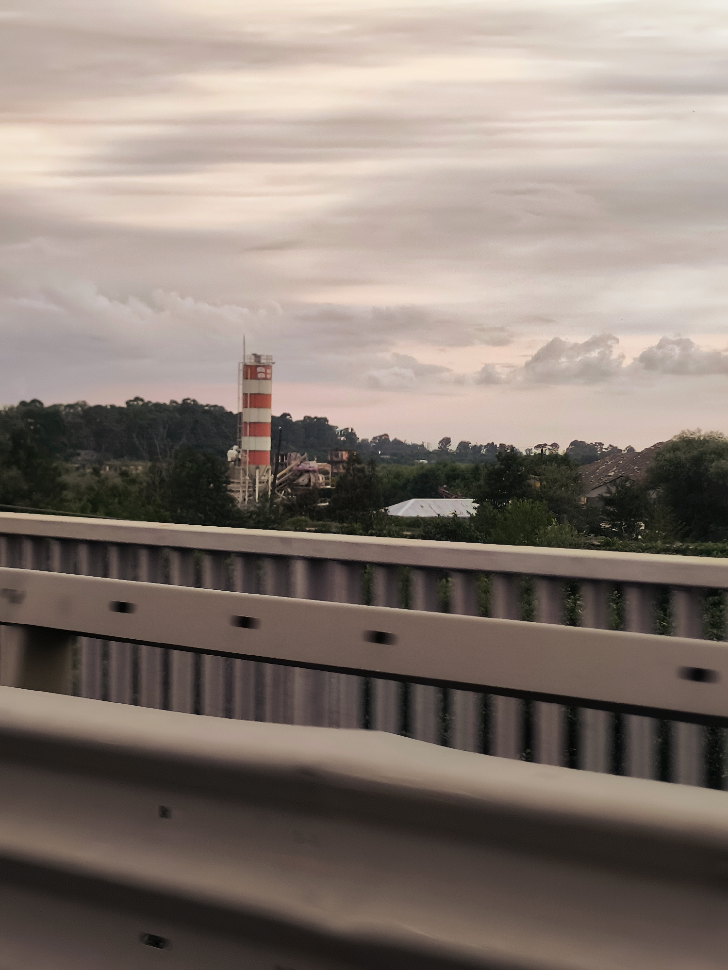 The image depicts a landscape scene with a distinctive red and white striped tower in the middle ground, possibly a silo or an industrial structure. The tower is situated amidst trees and other buildings, suggesting an industrial or semi-rural area. A white tarp-covered structure is visible to the right of the tower. In the foreground, a metal guardrail runs horizontally across the frame, indicating the viewpoint is from a road or elevated pathway. The sky is overcast with soft, muted colors, suggesting dusk or dawn. The overall atmosphere is calm and observational, with no people or explicit activity visible. The scene appears to be located in Charnali, Georgia, based on the provided information, though visual cues within the image itself do not specifically confirm this location.