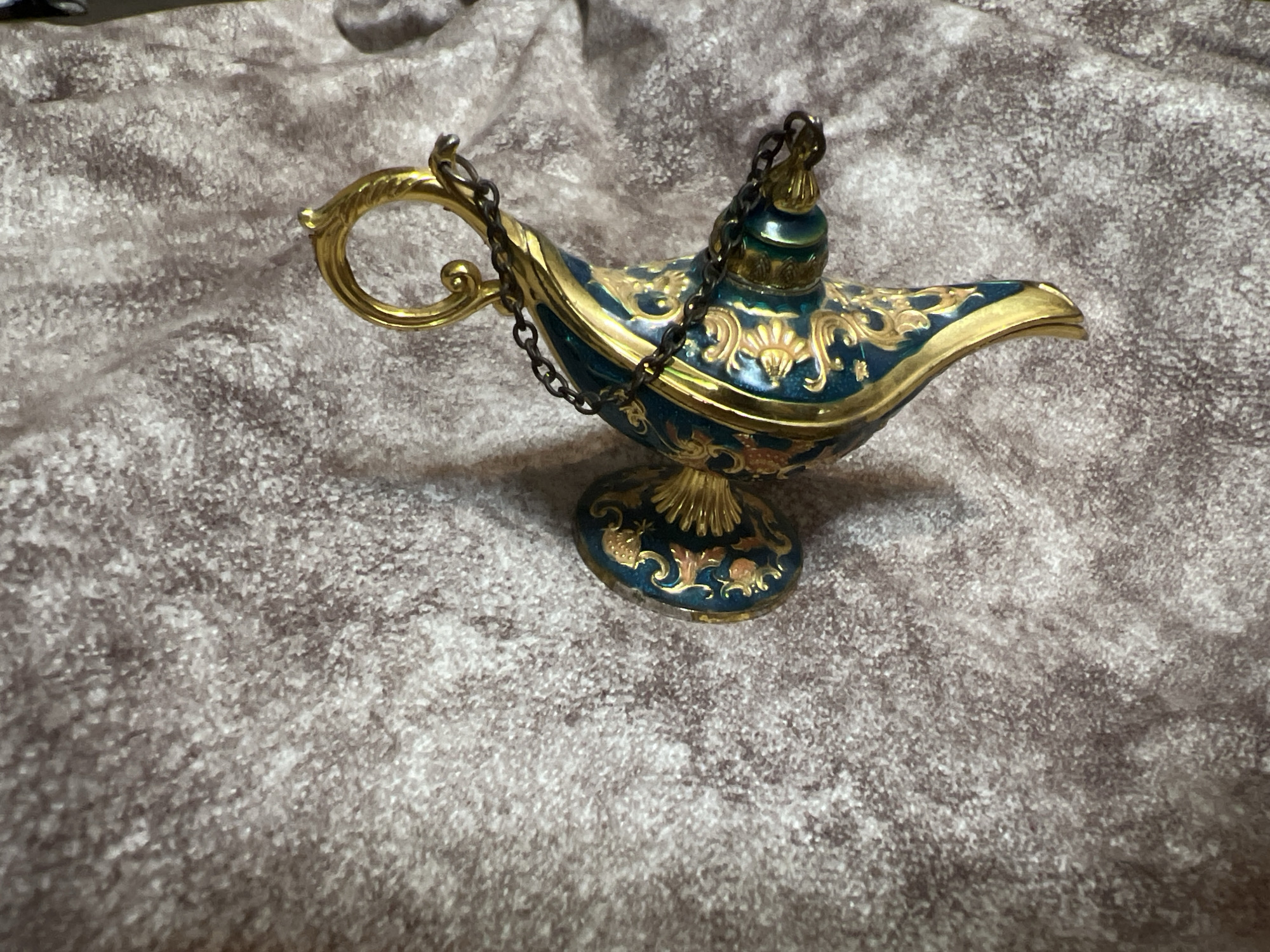 An ornate, antique-style magic lamp is positioned on a textured, greyish-brown fabric surface. The lamp is primarily a deep teal color with intricate gold embellishments and detailing. It features a curved handle, a spout, and a lid with a small chain connecting it to the handle. The gold patterns on the lamp appear floral and vine-like. The fabric surface beneath the lamp has a mottled, slightly fuzzy texture. There are no people or other objects visible in the image. The scene does not depict a location in Masazir, Azerbaijan.