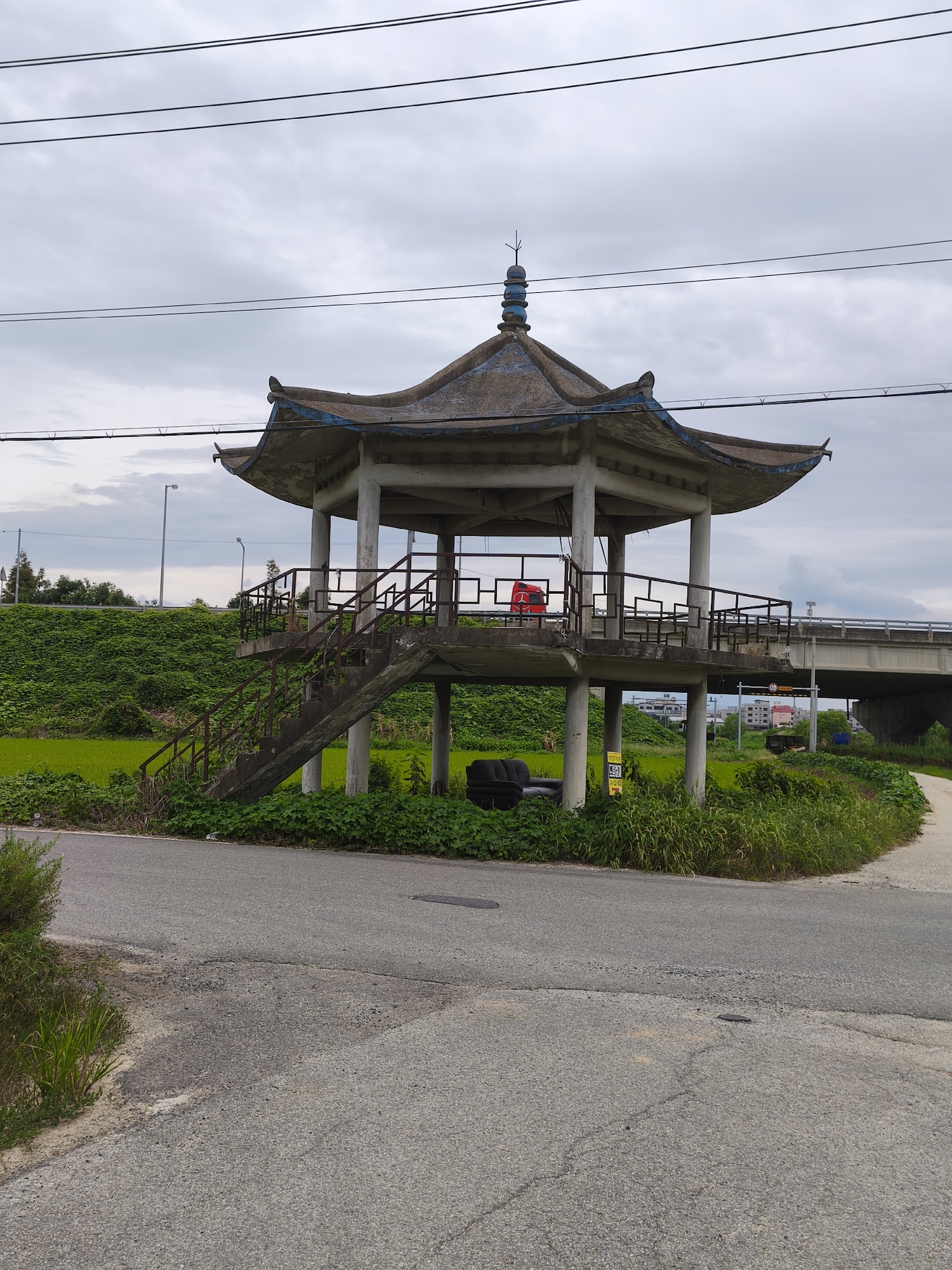 A concrete, octagonal pavilion with a tiered, pagoda-style roof is situated beside a rural road in Asan-si, South Korea. The pavilion is elevated on concrete pillars and accessible via a concrete staircase with rusting metal railings. Inside the pavilion, a red plastic chair is visible. At the base of the pavilion, two discarded tires and a black sofa are present, along with a yellow sign. The surrounding environment features lush green vegetation, including rice paddies, and a concrete overpass in the background. The sky is overcast. Power lines are strung across the upper portion of the image.