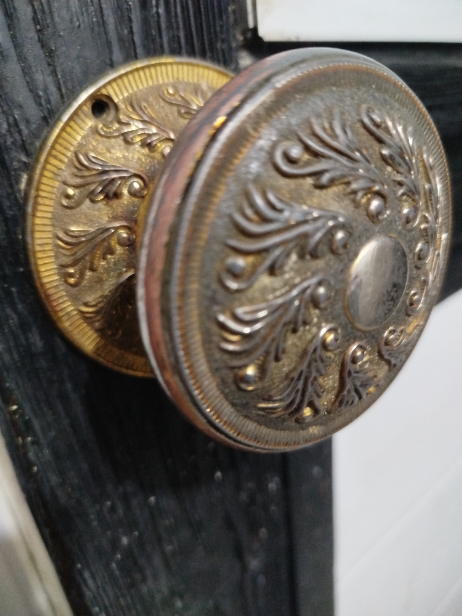A close-up, slightly angled shot shows a vintage, ornate doorknob on a dark, possibly wooden door. The doorknob is made of metal with decorative patterns, appearing to be brass on one side and a darker, possibly tarnished metal on the other. The left side of the doorknob is a circular plate with a raised, intricate floral pattern and a scalloped outer edge, finished in a golden hue. A small screw hole is visible on this plate. The right side of the doorknob is a larger, rounder knob with a similar decorative pattern, featuring raised swirling motifs and small raised dots, with a central circular indentation. This side has a more weathered, darker metallic appearance. The dark door has a textured, possibly wood grain surface, with faint highlights suggesting light reflection. The background to the right of the doorknob is a bright white, out-of-focus area, possibly a wall or another surface. The image lacks any discernible details about a specific city or location.