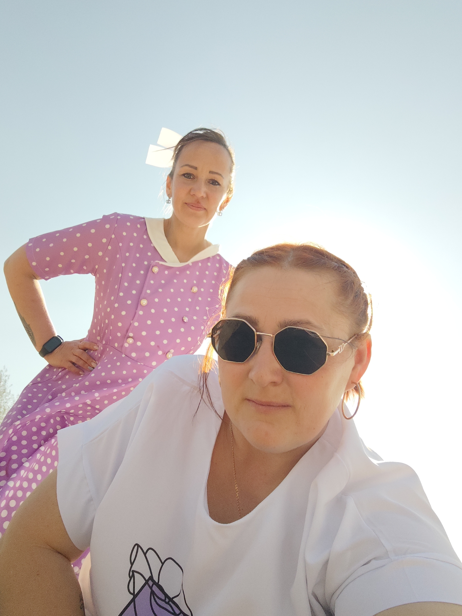 Two women are visible in an outdoor setting under a bright, sunlit sky. The foreground woman is closer to the camera and wears sunglasses, a white t-shirt with a graphic design, and hoop earrings. Her hair is pulled back. The woman in the background wears a vintage-style, polka-dot dress with a white collar and a white bow in her hair. She is standing further away, with one hand on her hip. The location is identified as Noviy, Russia.