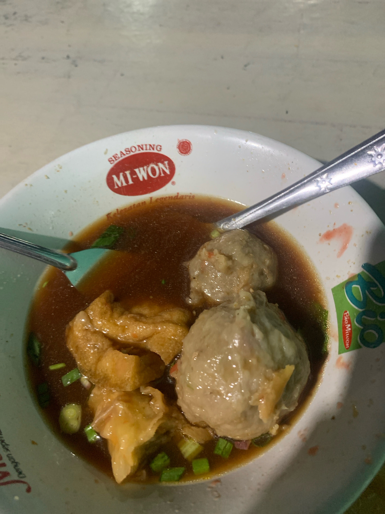 A close-up, overhead shot captures a bowl of bakso, a traditional Indonesian meatball soup, likely in the city of Tumpukrenteng, Indonesia. The white bowl, with a red and white "MI-WON SEASONING" logo on its rim, contains a rich, brown broth filled with several large, round meatballs, chunks of fried tofu, and chopped green onions. A silver spoon rests in the soup, its handle angled towards the top right of the frame, while a teal-colored spoon is partially submerged on the left. The surface the bowl sits on appears to be a light-colored, textured tabletop. The lighting is somewhat dim, casting subtle shadows and highlighting the glistening broth and textured surfaces of the food.