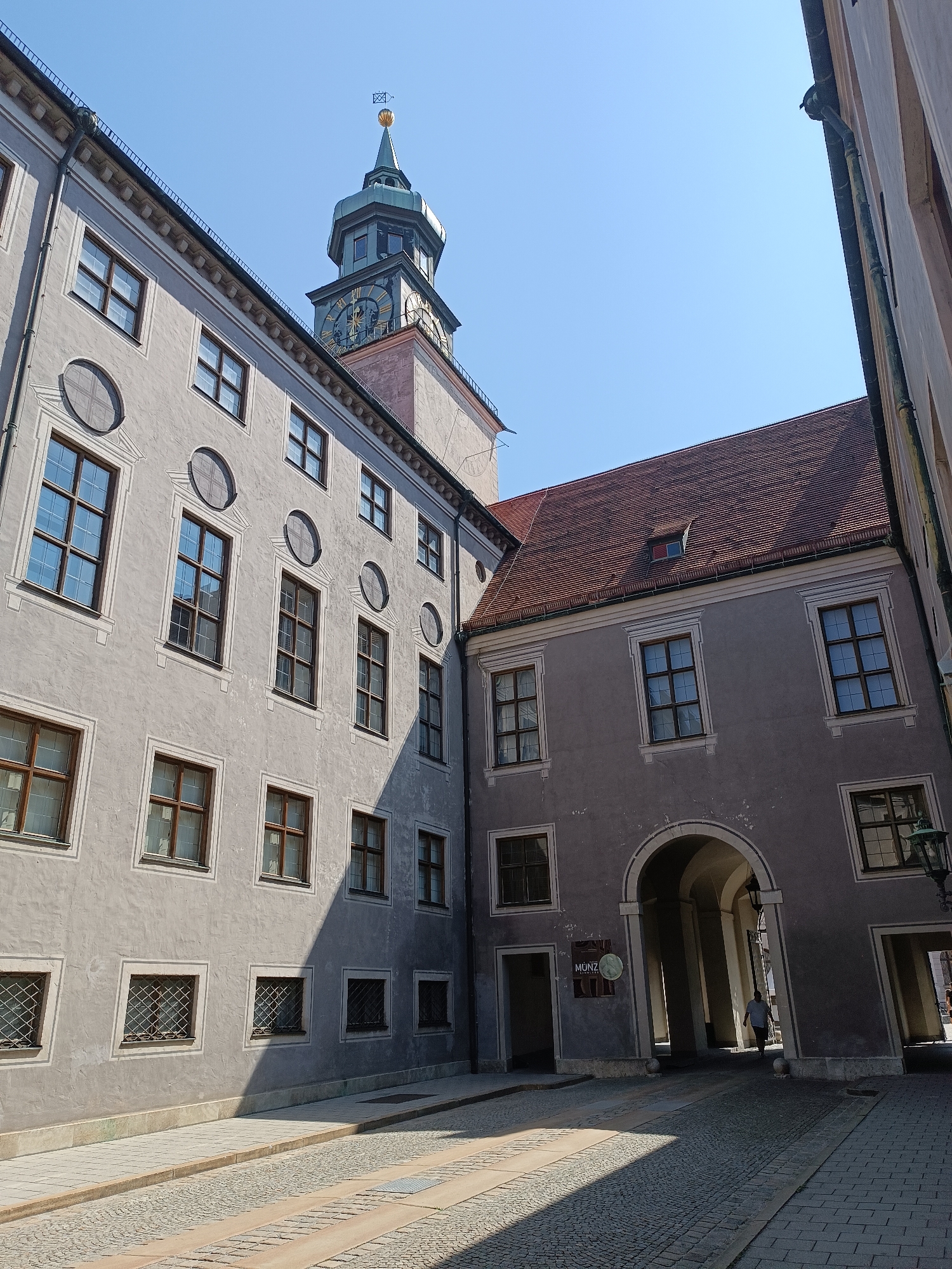 This is a photograph taken in Munich, Germany, likely of the Münzamt building, part of the Residenz complex. The image features a courtyard area with historic architecture and a clear blue sky.
In the foreground, a cobblestone street curves to the right, with a paved walkway along the left side of the courtyard. The street is cast with strong shadows from the surrounding buildings, indicating a bright sunny day.
The dominant architectural elements are the grey-washed facades of multi-story buildings. The building on the left has numerous windows, some with decorative surrounds. Circular decorative elements are also visible on this facade. Towards the top of this building, a portion of a clock tower with a green spire and golden weather vane is visible against the sky. The clock face has roman numerals and ornate hands.
The building on the right features a large arched entryway supported by columns, leading into a darkened interior passage. Several windows are visible on this facade as well, including a small dormer window on the red-tiled roof. A traditional lantern hangs from the facade near the archway.
A single person, wearing a light-colored shirt and dark pants, is seen walking through the arched entryway. Another person is partially visible in the far right background, also appearing to walk away from the camera. A sign with the word "MÜNZ" is affixed to the wall near the archway.
The overall impression is a serene, sun-drenched scene within a historic urban setting.
