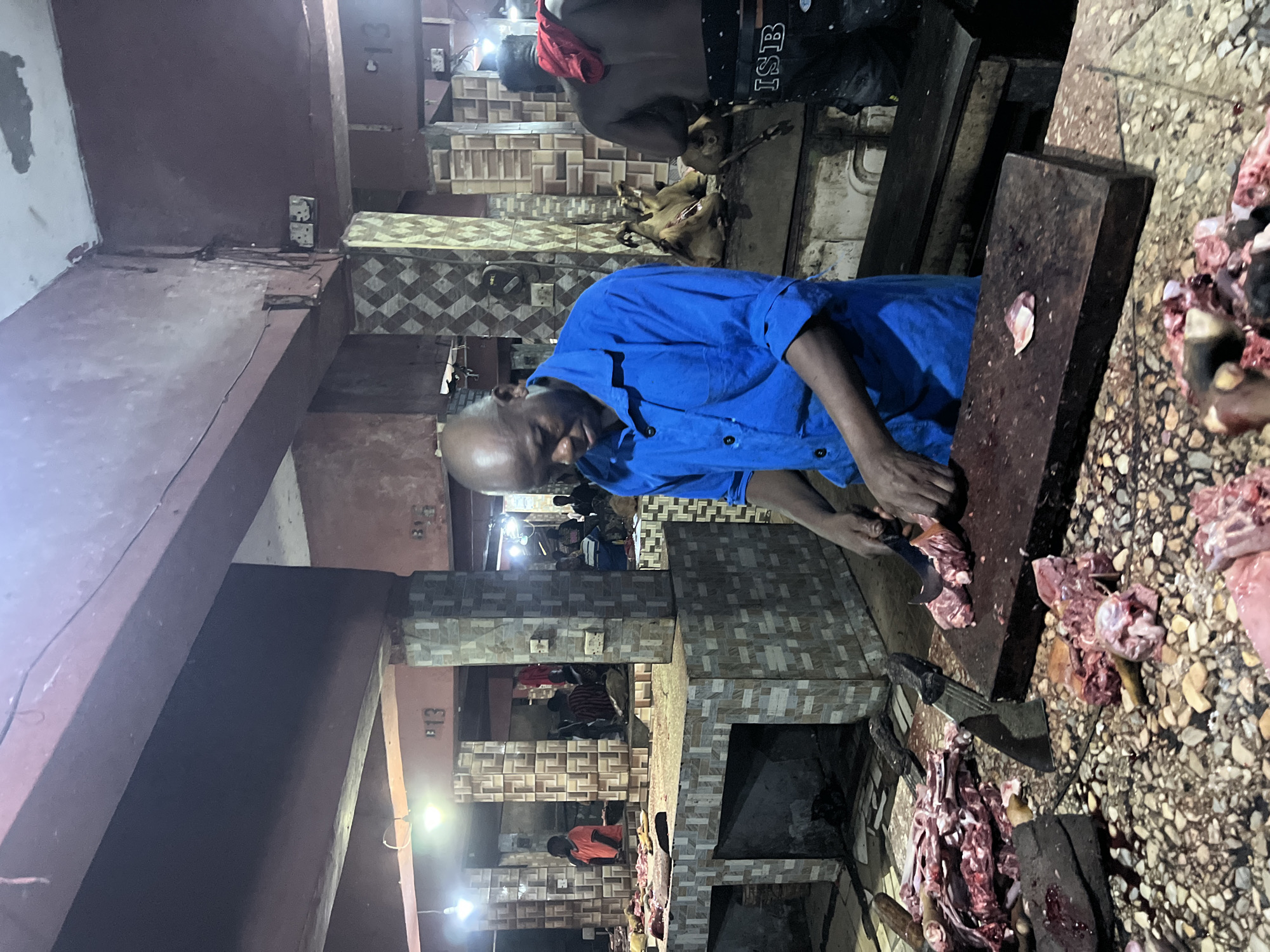 The image depicts a bustling butcher shop, likely located in Nkpor, Nigeria. The foreground is dominated by a rough, speckled countertop covered with raw meat, some of which appears to be in pieces and prepared for sale. A large, dark wooden cutting board rests on the counter, with a cleaver and another knife nearby. In the center of the image, a bald Black man wearing a bright blue collared shirt is actively engaged in butchering meat with a knife. His gaze is directed downwards at his task. To his right, another Black man, partially visible, is also working, with a red cloth draped over his shoulder. The background reveals more butchering stalls with similar countertops, where other individuals can be seen attending to customers or preparing meat. The architecture suggests an indoor market setting with exposed beams and tiled pillars. Lighting appears to be artificial, with overhead bulbs illuminating the scene. The overall atmosphere is one of active commerce and food preparation.