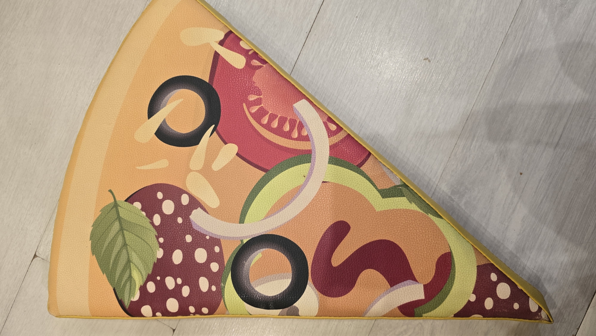 A close-up, overhead, indoor shot shows a triangular-shaped pillow designed to look like a slice of pizza, lying on a light-colored wooden floor. The pillow has a yellow edge and features a stylized representation of pizza toppings, including what appears to be pepperoni slices with white dots, black olives, red tomato slices, green bell pepper rings, and a basil leaf on a tan and orange background representing the pizza crust and cheese. The lighting is neutral and diffused, casting no distinct shadows. The environment is an indoor setting, likely a room with a floor made of light-colored wood planks. There are no people or animals present in the image. The exact location in the city of Chaikovsky, Russia, is not discernible from the visual information provided.