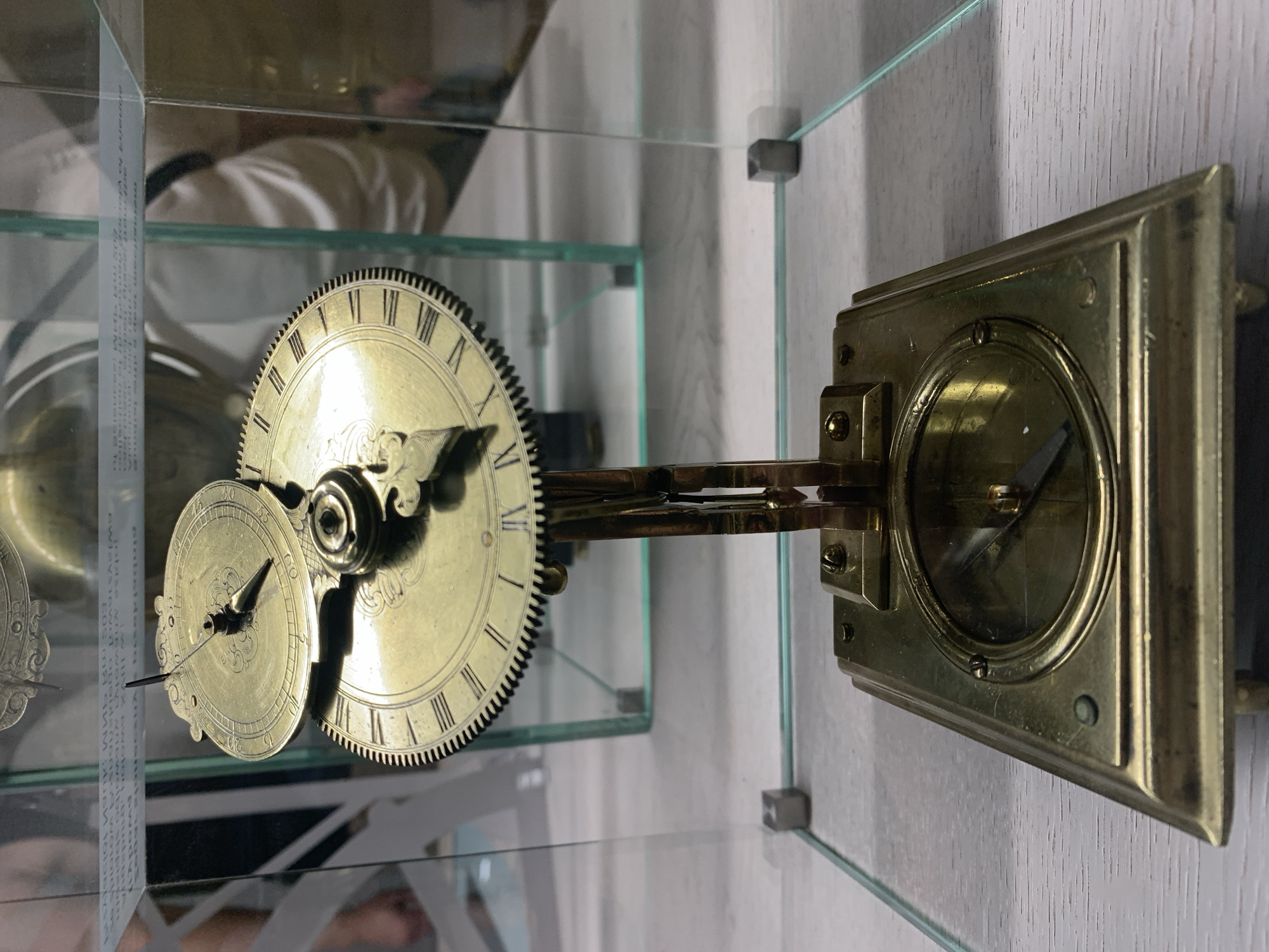 This image shows a close-up view of an antique brass clock, likely a horological artifact, displayed within a glass case. The clock features multiple dials and intricate gear mechanisms, with Roman numerals visible on at least one of the faces. The clock is supported by a brass stand and rests on a base that contains a circular display, possibly another gauge or dial. The background is blurred, but appears to be an indoor setting with reflections of people and exhibits in the glass enclosure, suggesting it is part of a museum or exhibition.
The horological artifact is located at the National Museum in Poznan, Poland, in the Department of Decorative Arts. This specific artifact is identified as a "Regulator with pendulum and escapement of a unique design, late 18th century," attributed to Jan Chrzciciel Zydlitser. The regulator, known for its precision in timekeeping, is a significant piece within the museum's collection, showcasing the advanced craftsmanship of the period and the history of horology.