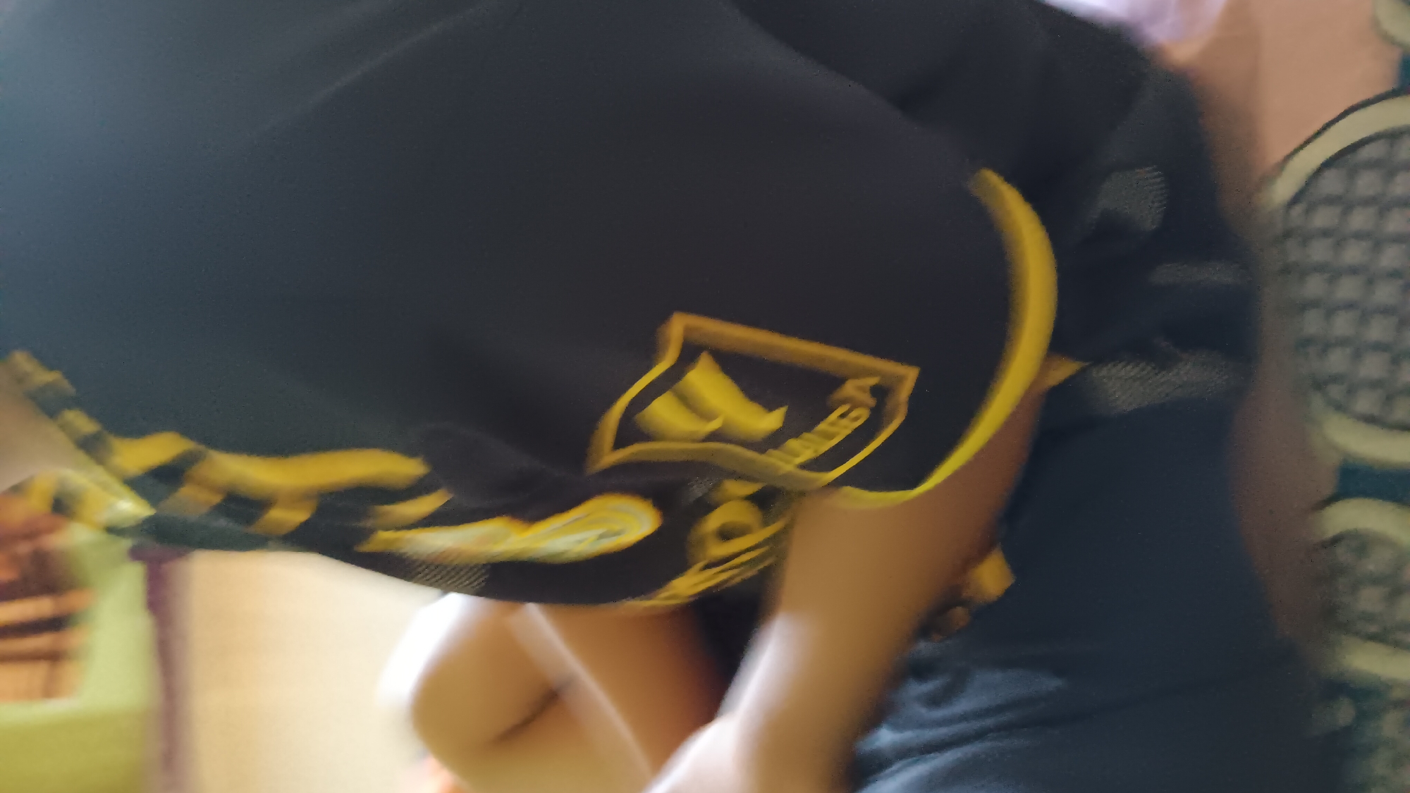 The image shows a close-up of a person's torso and legs, wearing a dark blue shirt with yellow accents and lettering on the sleeve. The person is sitting or lying down, with their legs bent. The background is blurred but appears to be an indoor setting with bedding or upholstery. There are no identifiable landmarks or environmental clues that would suggest a specific location within Sonobekel, Indonesia. The image is too blurry and lacks detail to provide any further specific information about the scene or its inhabitants.