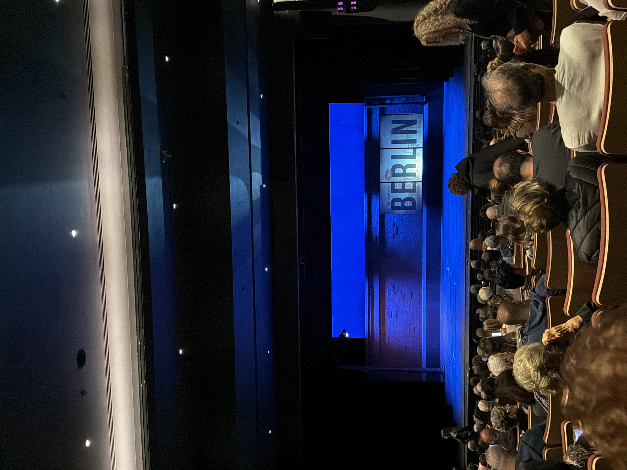 The image shows a theater audience sitting in rows of seats facing a stage. On the stage, there is a backdrop with the word "BERLIN" written vertically in large letters. The stage is lit with blue light. The audience members are facing the stage, and some are looking at their phones. The location is a theater, but not in Tel-Aviv, Israel.