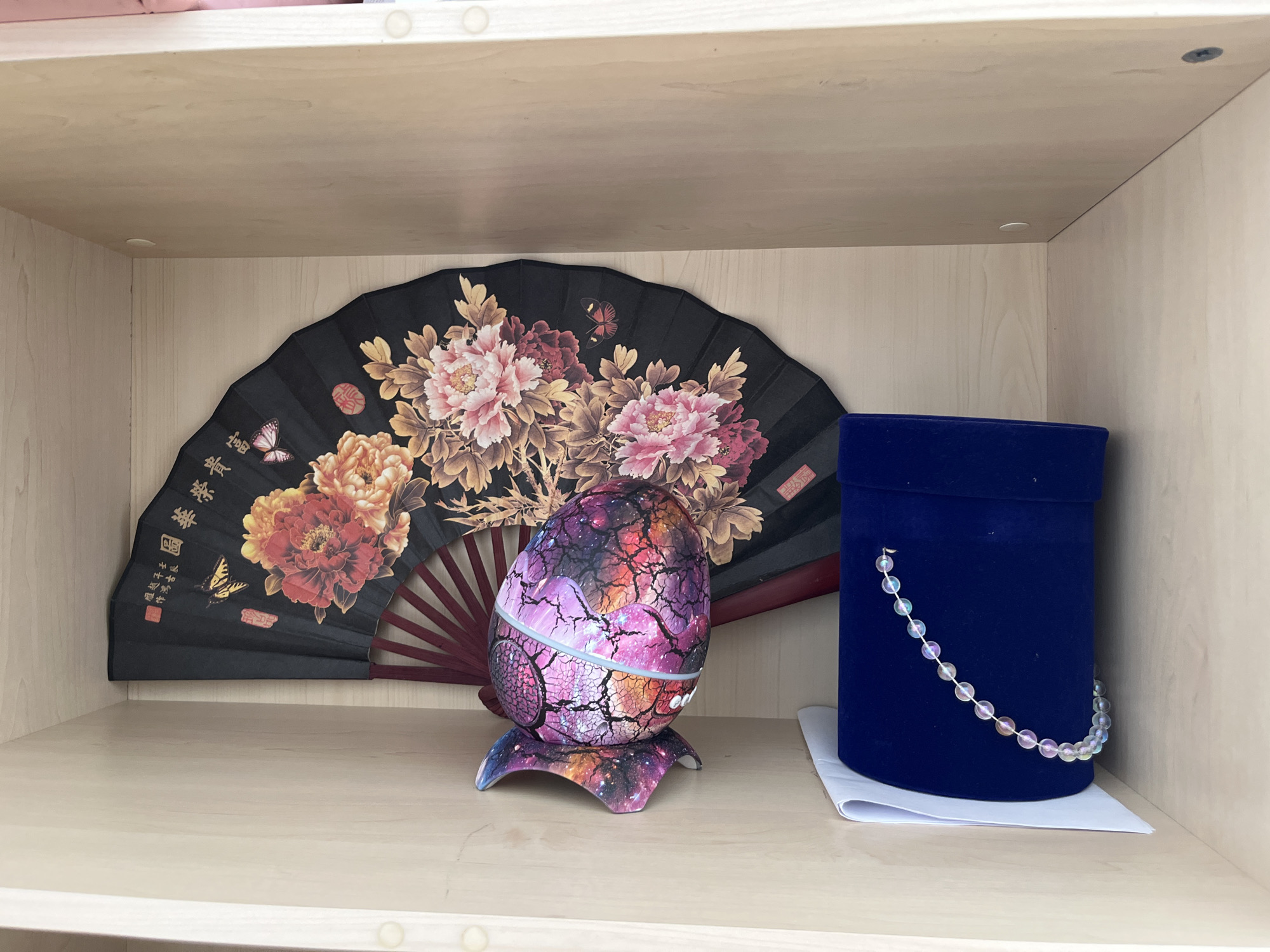 An open wooden shelf displays three decorative items. On the left, a black fan is open, revealing a detailed painting of pink and orange peonies and butterflies. In the center, an egg-shaped object with a galaxy-like pattern sits on a matching stand. To the right, a small, cylindrical dark blue velvet box is placed on a white sheet of paper. A strand of iridescent beads drapes over the front of the box. The shelf is made of light-colored wood, and the background consists of vertical wooden planks.