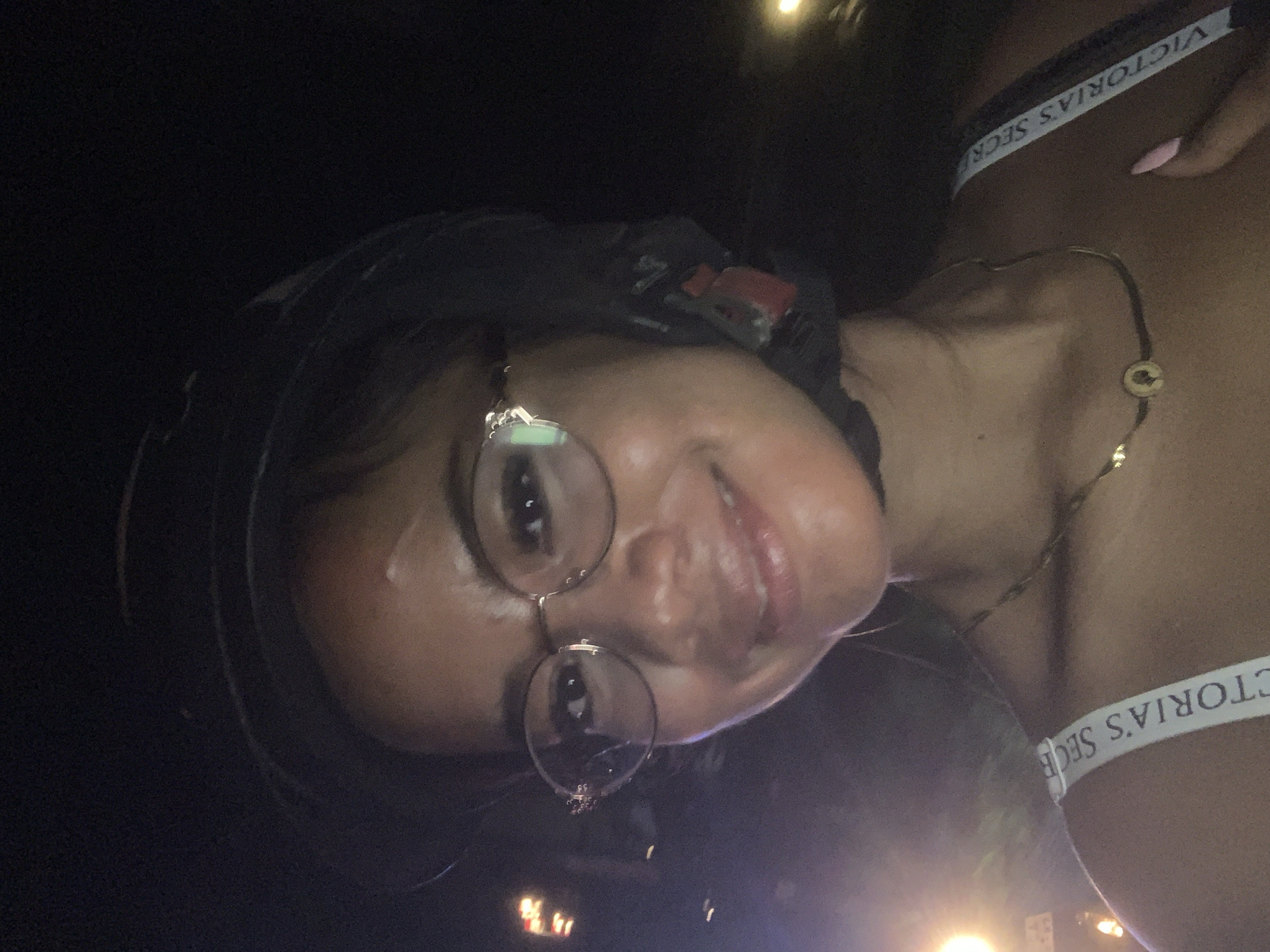 A woman with brown skin, wearing round glasses and a black helmet, is smiling. She is wearing a necklace and a white bra strap with the words "VICTORIA'S SECRET" on it. The location may be North Kuta, Indonesia, though this is not visually verifiable.