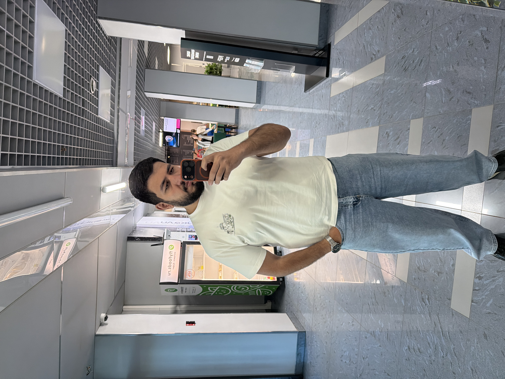 A man is taking a selfie in a modern, brightly lit building. The building has a gray, tiled floor with white rectangular inlays. The walls are a mix of gray and white, with a grid-like pattern on one side and a series of glass-fronted cabinets on the other. One of the cabinets has branding on it that indicates it may be refrigerated. A black display unit stands nearby. The man is wearing a white T-shirt, blue jeans, and a watch. The location may be related to an airport transit terminal, based on the presence of retail units. Krasnoyarsk, Russia, is referenced via some text.