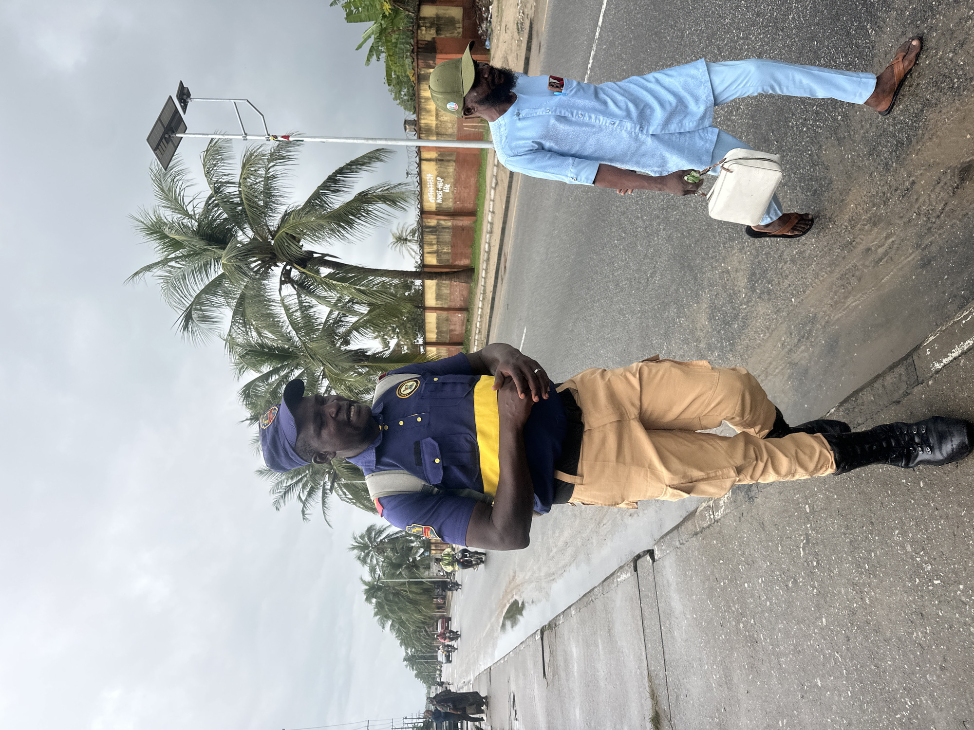 In Badagry, Nigeria, two men walk along a rain-slicked asphalt road under a cloudy sky, with palm trees lining the left side of the road. The man on the left, closer to the foreground, wears a dark blue uniform with yellow stripes across the chest and a matching blue cap with an emblem. He also sports tan cargo pants and black boots, and carries a light-colored backpack. The man on the right wears a light blue traditional African tunic and pants, a green cap, and brown sandals, and carries a white rectangular bag in his right hand. In the background, a street lined with palm trees, a solar-powered streetlight, and a low wall with some signs are visible. Several motorcycles and a few cars are seen on the road, with more people walking on the sidewalk to the far left. The overall scene depicts a street in Nigeria on a possibly rainy day.