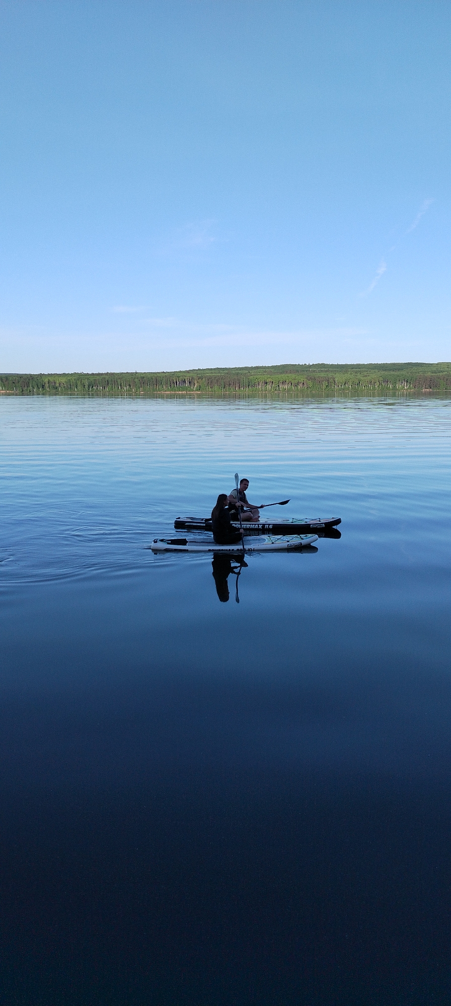 The image depicts two individuals on a paddle board in a body of water, likely a lake or river. The paddle board is labeled "POWERMAX 11.6". One person is sitting and holding a paddle. Their reflections are visible on the water surface. The background shows a treeline, and the sky is clear and blue. The specific location is in the city of Chaikovsky, Russia.