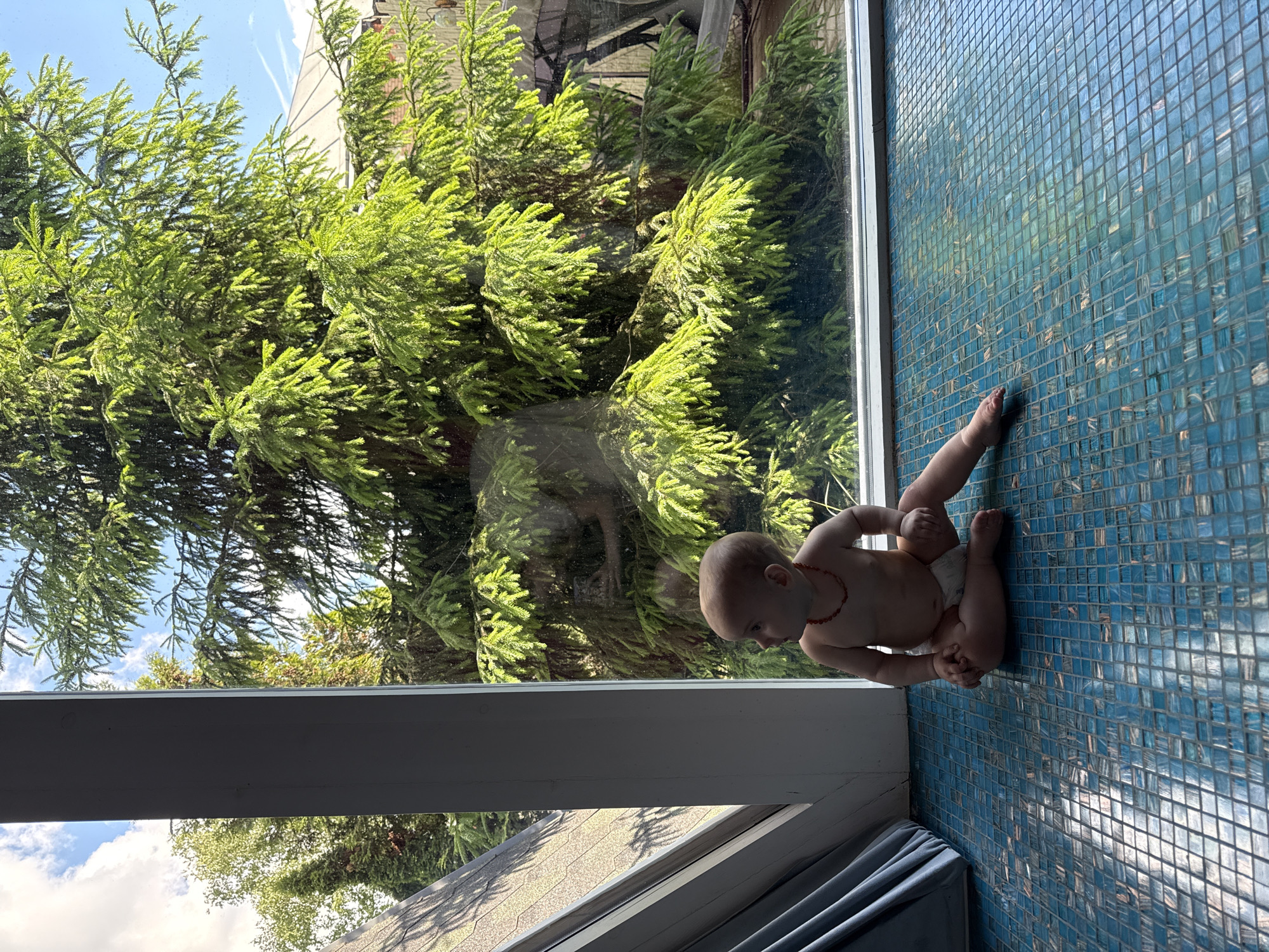 A partially clothed baby sits on a blue-tiled surface near a window. The window shows a dense green tree and sky. The location is likely indoors. The city of Чёрное, Russia, cannot be verified from this image.