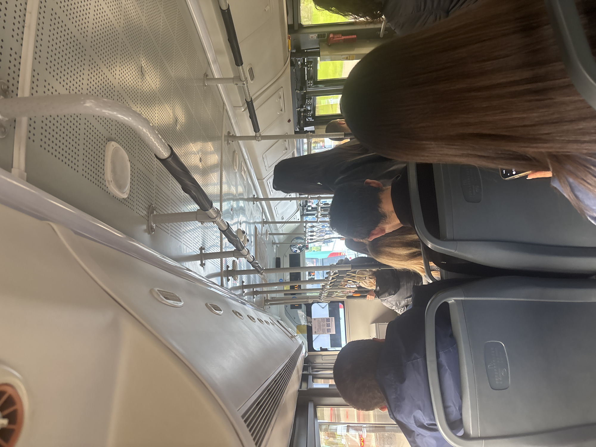 The image depicts the interior of a bus, presumably in Yerevan, Armenia. Passengers are seated in rows facing forward. The interior of the bus is white and grey, with visible ventilation systems and handrails. The bus appears to be in transit, as indicated by the outside scenery visible through the windows. There is no definitive evidence within the image to conclusively confirm that the location is Yerevan, Armenia.