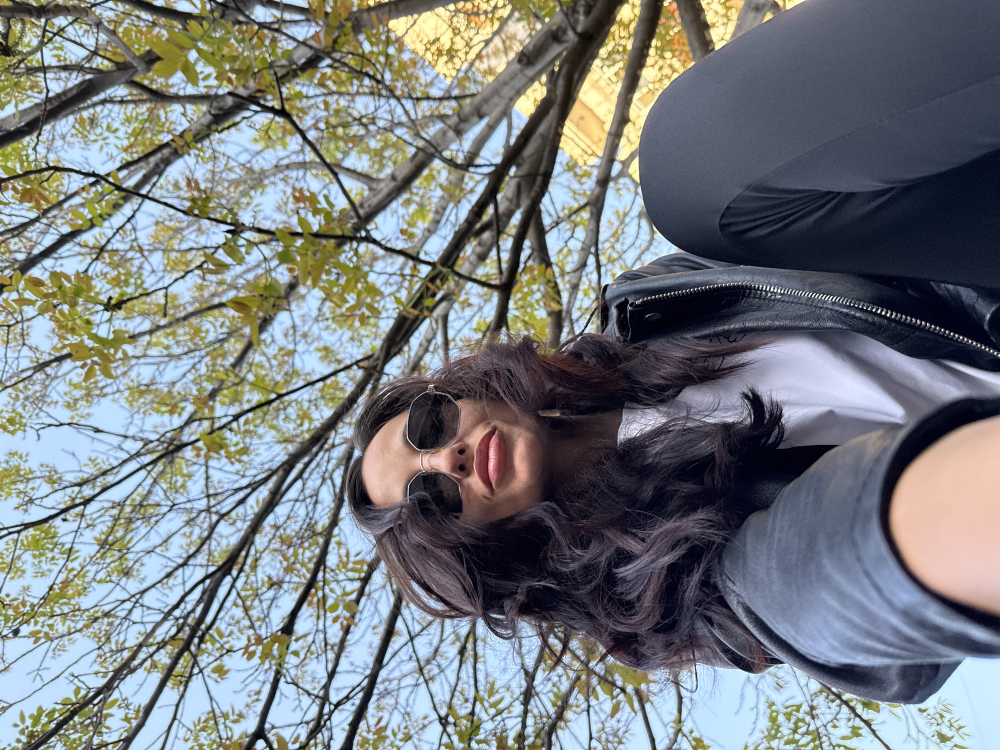 A low-angle, upward-facing photograph depicts a woman with dark, wavy hair and sunglasses, wearing a white shirt and a dark leather jacket. Part of her dark pants are visible in the upper right. The backdrop is a sky with light green tree branches with yellowish leaves. The location is potentially Pleven, Bulgaria.