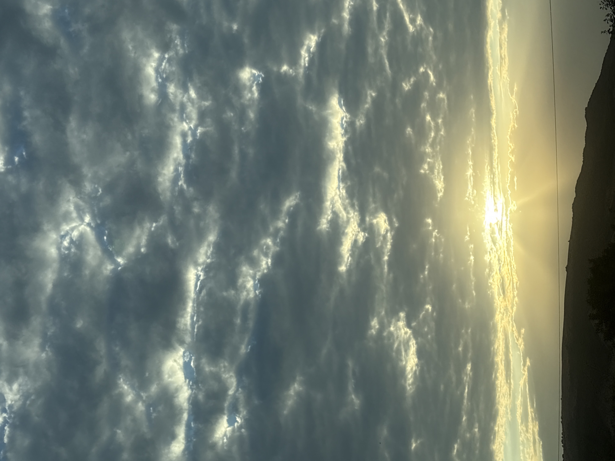The image shows a cloudy sky with the sun partially visible behind a band of clouds, casting rays of light downwards. Below the sky, a dark silhouette of a hilly landscape can be seen, with a thin horizontal line that might be a power line or a fence. The image appears to be taken outdoors, possibly in the rural outskirts of Lysogorskaya, Russia, given the hilly terrain and the vast expanse of sky. There are no people or specific objects clearly identifiable in the foreground or midground, making the focus solely on the atmospheric conditions and the landscape.