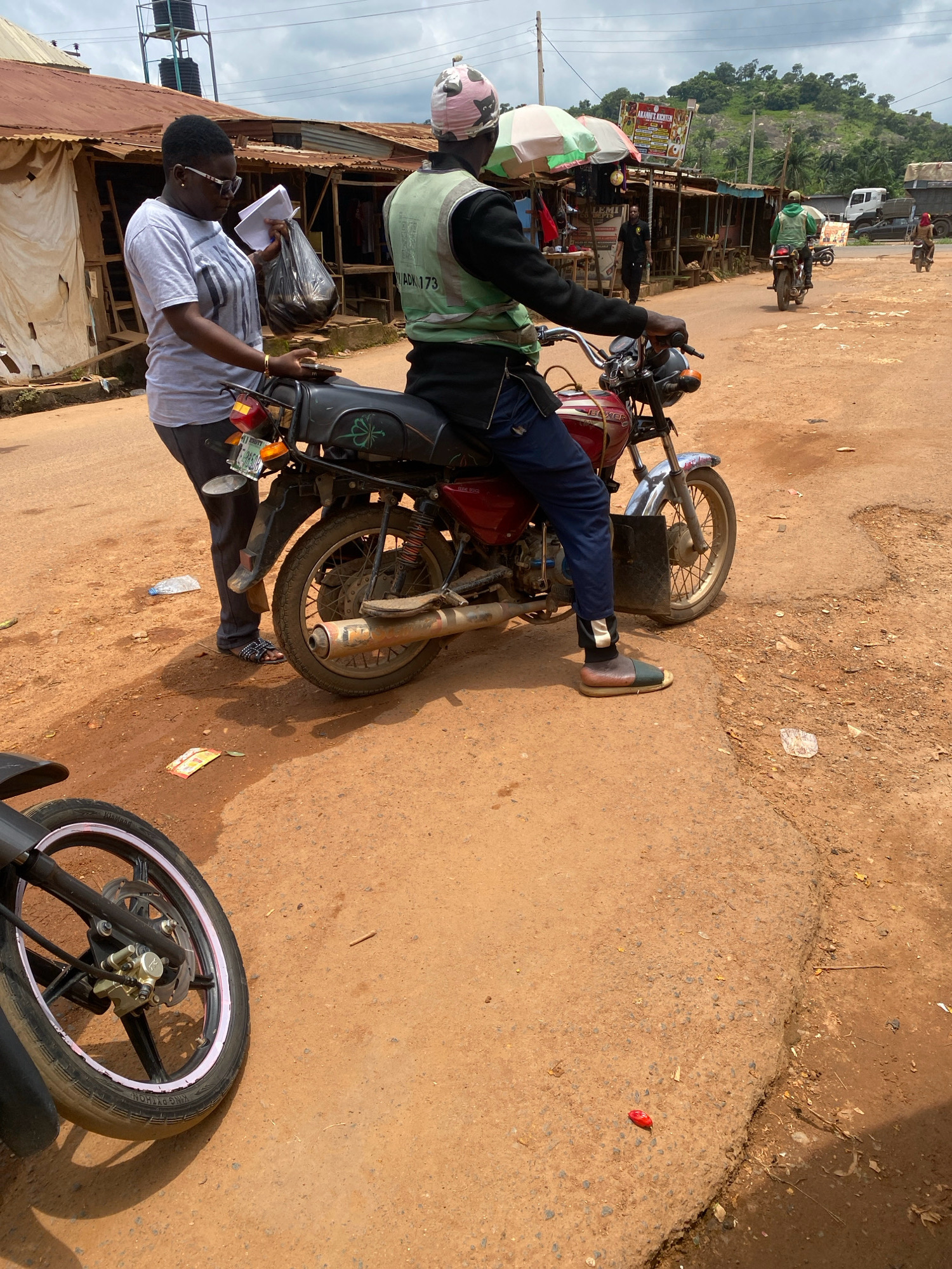 In Iworoko, Nigeria, a woman stands next to a motorcycle, holding a plastic bag and papers. She is wearing a grey t-shirt, dark trousers, and sandals. The man on the motorcycle is wearing a green vest with "ADV.173" visible on it, a patterned head covering, and dark trousers. The motorcycle is red and black. In the background, there are several market stalls with corrugated iron roofs and white fabric coverings. Beyond the stalls, a hilly, vegetated landscape is visible under a cloudy sky. Several other motorcycles and vehicles are present in the distance. The ground is a mix of dirt and gravel.