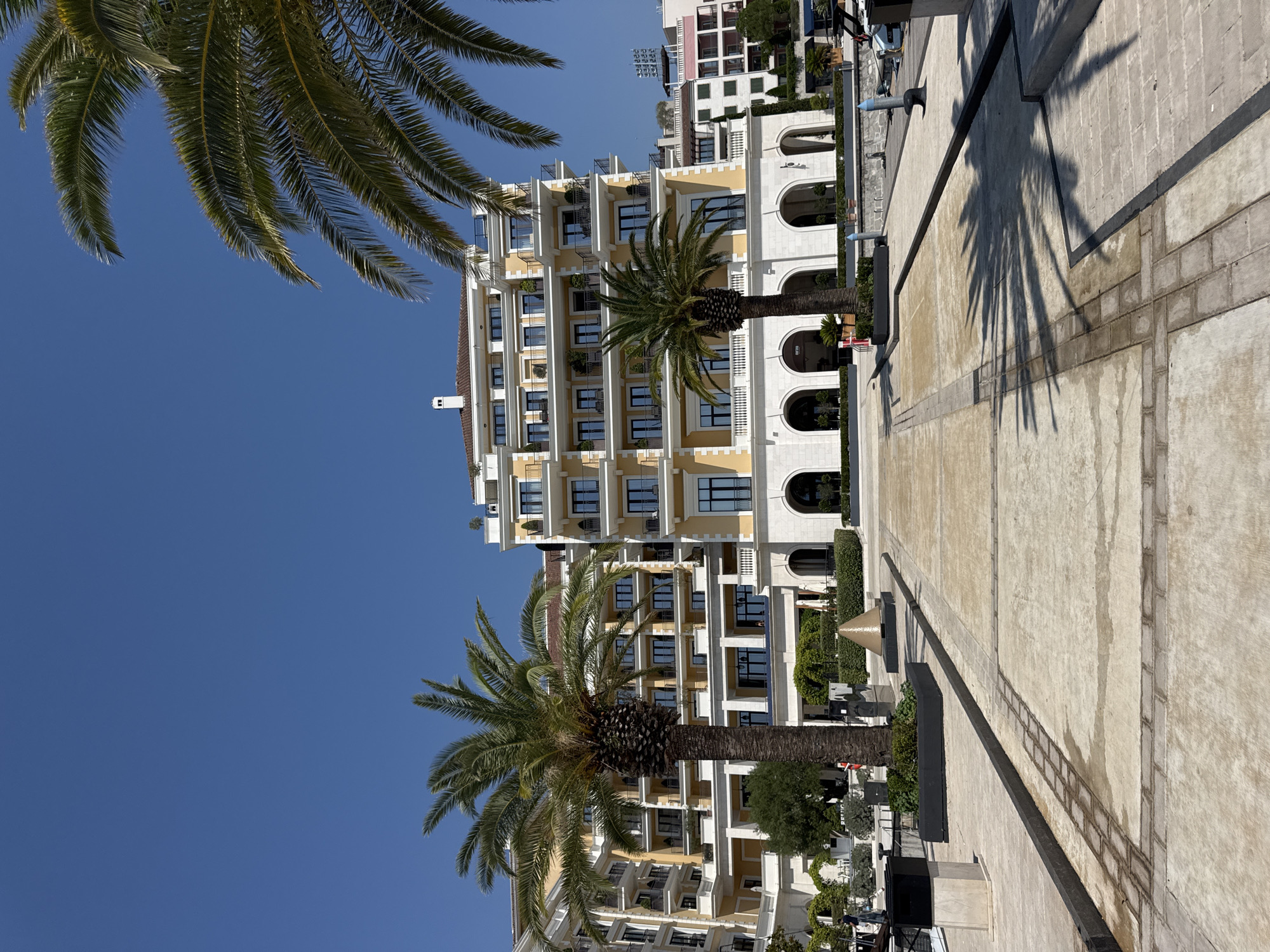 In Tivat, Montenegro, a bright, sunny day showcases a modern hotel with a yellow facade and multiple balconies, set against a clear blue sky. Two prominent palm trees stand in the foreground, their fronds casting shadows on the paved walkway. The walkway is made of concrete and stone pavers, with a dark border running along its length. In the background, other buildings and greenery are visible, suggesting a resort or waterfront area. The scene depicts a tranquil outdoor environment, likely a public space within the hotel complex or adjacent to it.