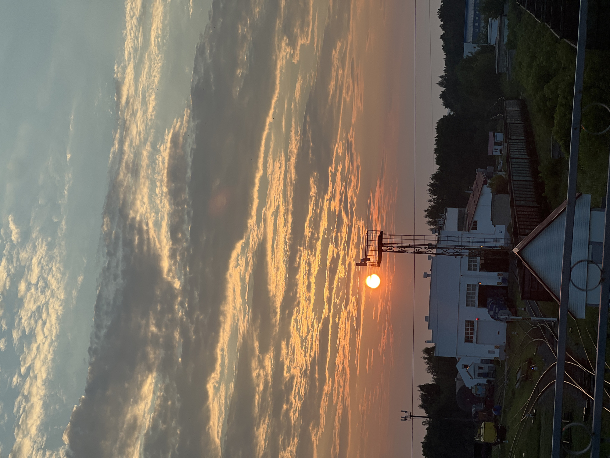 This photograph captures a sunrise over a train depot, likely located in Chaikovsky, Russia. The sun, a brilliant orange orb, is low on the horizon, casting a warm glow across a sky filled with striated clouds. The clouds are illuminated in shades of orange, yellow, and gray, with hints of blue peeking through.
In the foreground, a complex network of train tracks curves and converges. Several train cars, including freight cars and what appear to be maintenance vehicles, are visible on the tracks. Prominent structures include a large, white building with multiple garage-like doors, possibly a maintenance shed or depot. Adjacent to it is a tall, metallic tower with a platform at its top, likely for signaling or observation. Other smaller buildings and a densely wooded area are also present in the background, suggesting a semi-industrial or peri-urban setting. The overall atmosphere is one of the early morning hours, with the industrial landscape bathed in the soft, ethereal light of dawn. No people are clearly discernible in the image.