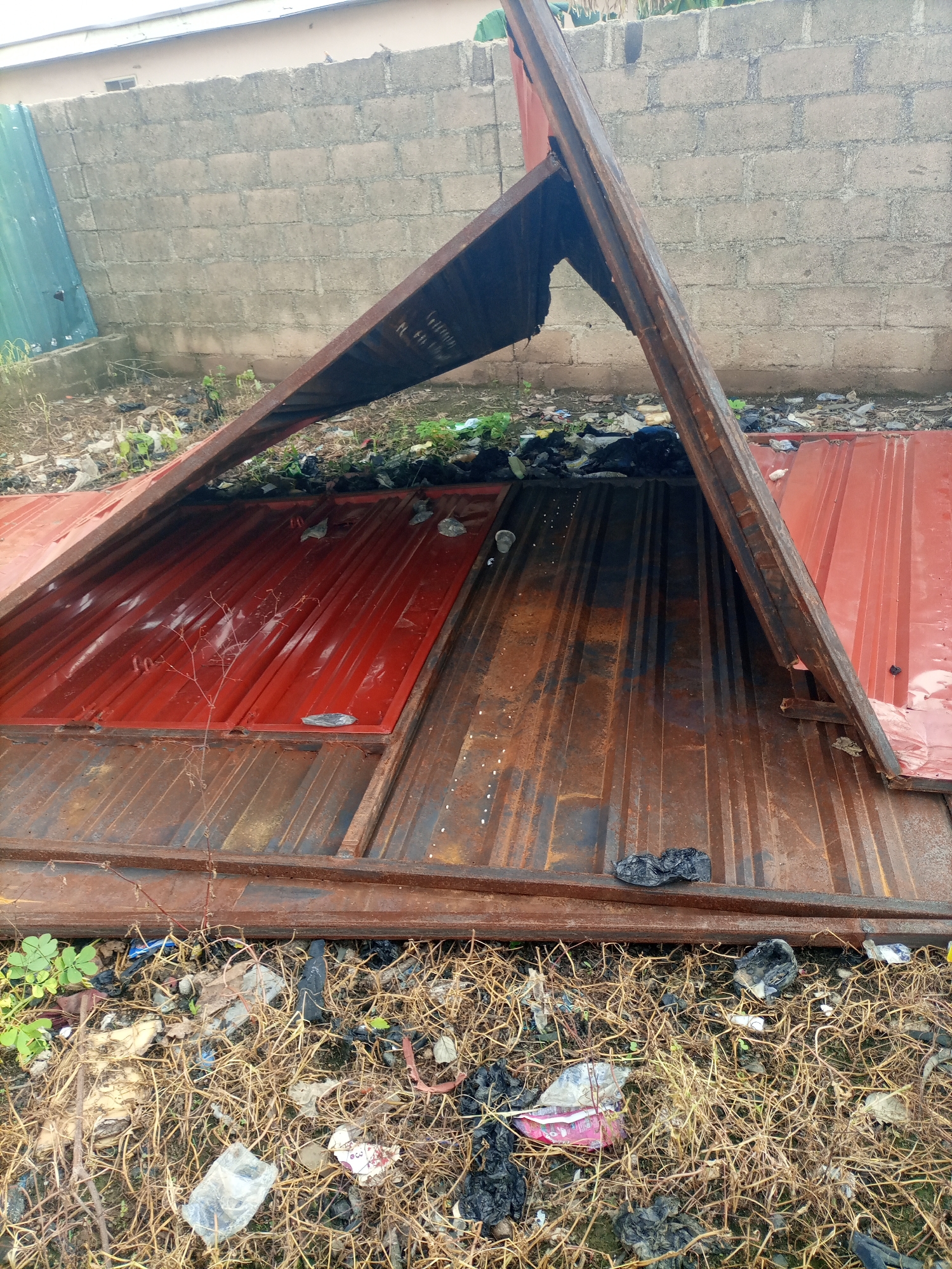 The image depicts a scene of debris and damaged corrugated metal roofing sheets, possibly the aftermath of a fire, located in Jalingo, Nigeria. The roofing sheets, some red and some rusted, are bent and broken, forming an inverted 'V' shape in the center of the frame. They lie on the ground amidst dry grass, twigs, and a significant amount of scattered trash, including plastic bags, wrappers, and what appear to be charred debris. In the background, a wall constructed of concrete blocks is visible, with a portion of a green metal structure to the left. The overall environment suggests an unkempt and neglected outdoor space within the city. There are no people or animals present in the image, and no actions or interactions are observable.