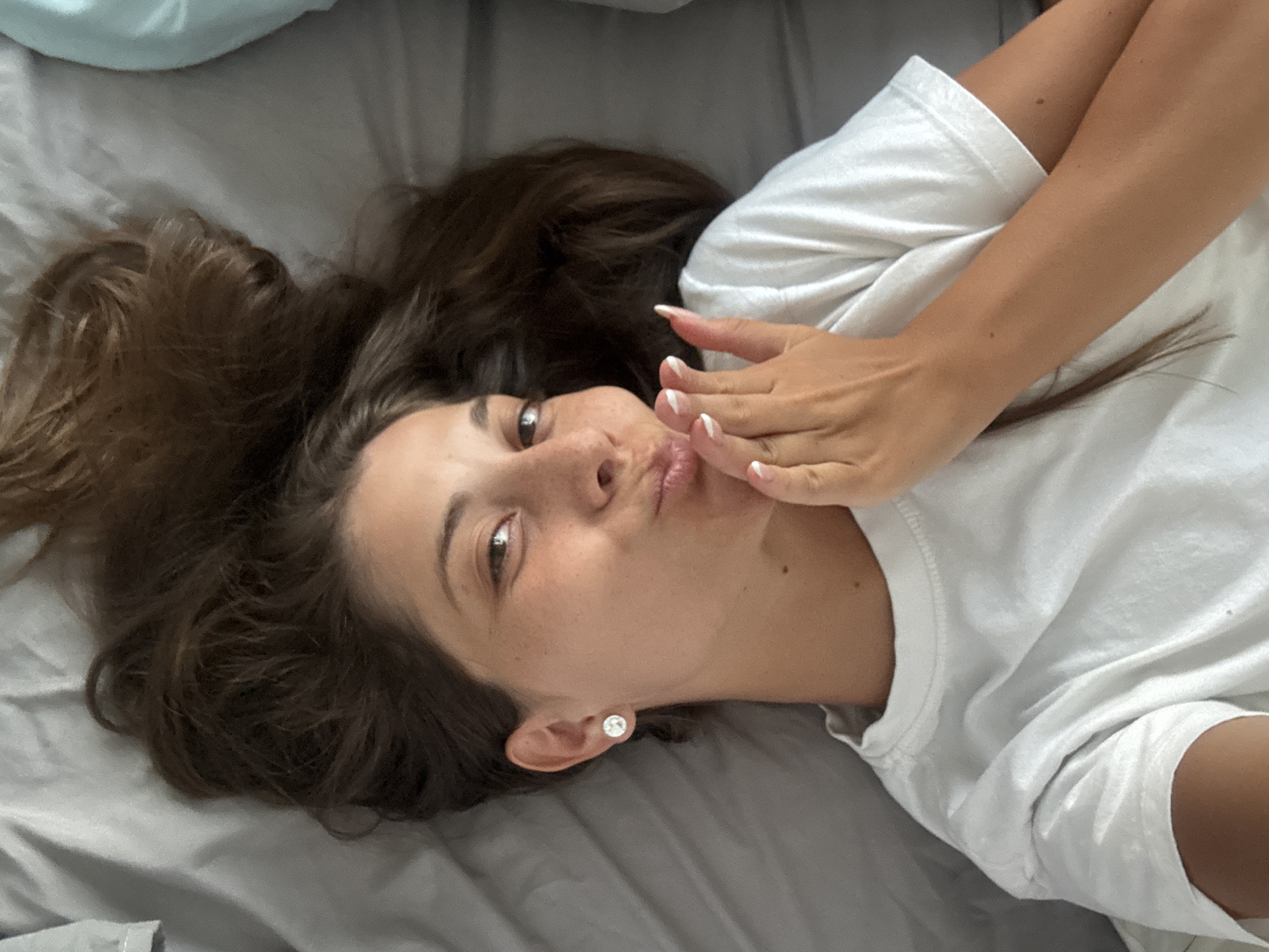 A woman is lying on a bed. She has long, brown hair and fair skin with visible freckles. She is wearing a white T-shirt and a single stud earring in her left ear. Her left hand is raised to her face, near her mouth, with fingers slightly curled. Her nails are manicured with a French tip. The bedding appears to be a gray, textured fabric. The background is out of focus, but the edge of a blue pillow is visible. The image provides no information about location in Đà Nẵng, Vietnam or any other geographical data.
