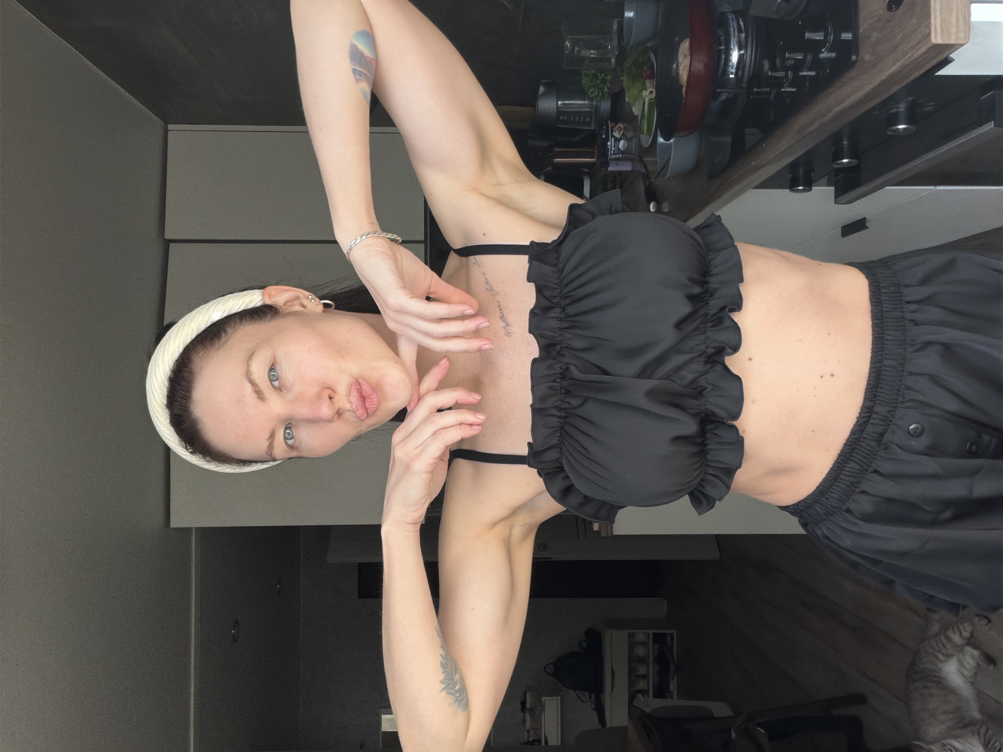 A woman with light eyes, a white headband, and two arm tattoos poses indoors. She is wearing a black, ruffled crop top and black pants with buttons. Her right arm is above her head, and her left arm is near her face. Behind her are kitchen cabinets, countertops with kitchenware, and a partial view of a room with a cat. The location is likely Perm, Russia, based on metadata or other identifying information not explicitly provided in the image.