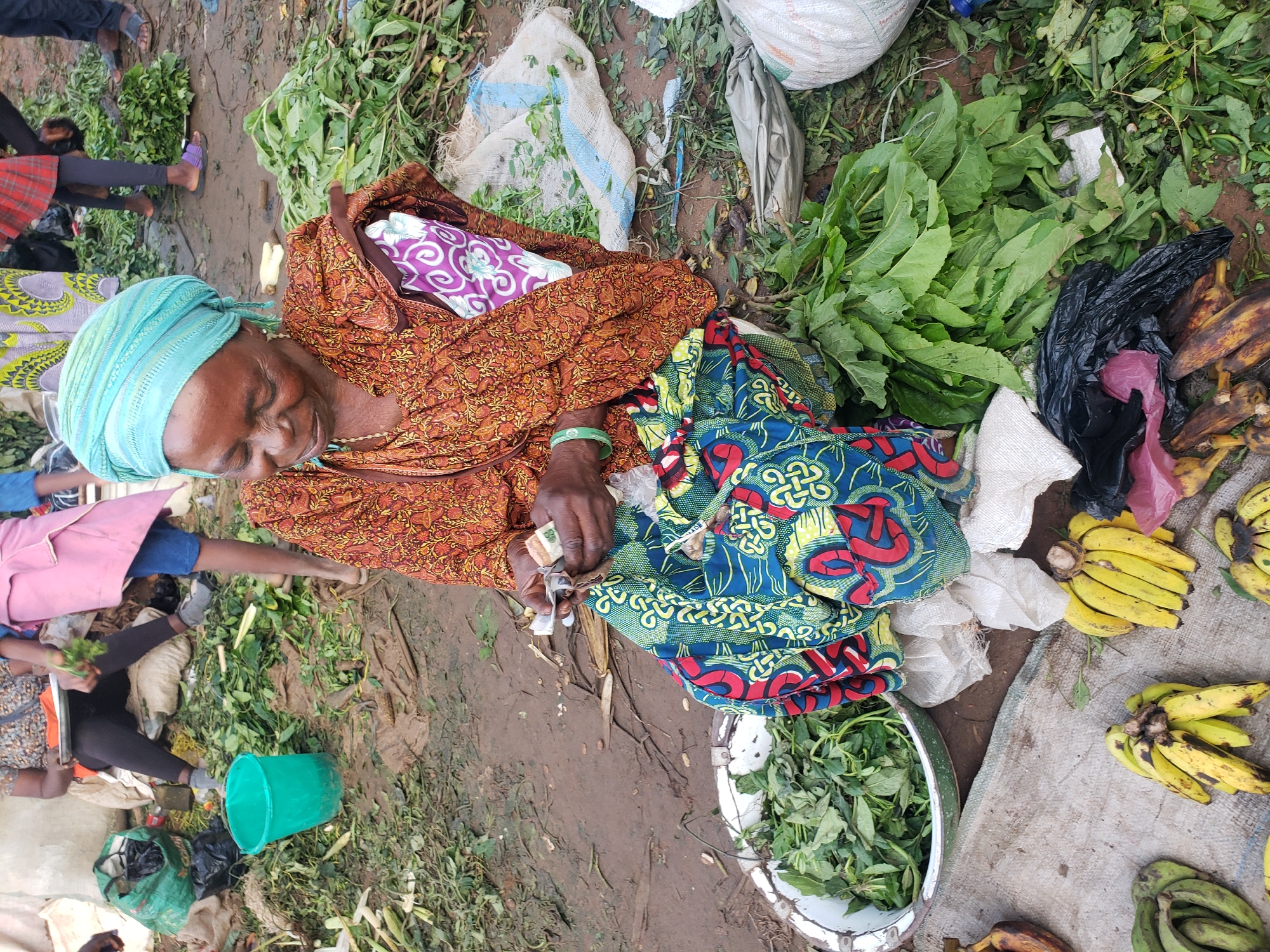 In Kakuri Gwari, Nigeria, a woman wearing a blue headwrap and an orange patterned dress sits on the ground amidst piles of green leafy vegetables and produce. She is counting money in her hands. To her left, a green bucket sits near a pile of similar vegetables. In front of her, a large bowl overflows with leafy greens. Various other produce, including bananas and what appear to be roots, are scattered around the scene. Bags and sacks, some filled with produce and others empty, are also present. The ground is dirt and appears damp. In the background, other individuals are partially visible, some also surrounded by produce and bags.